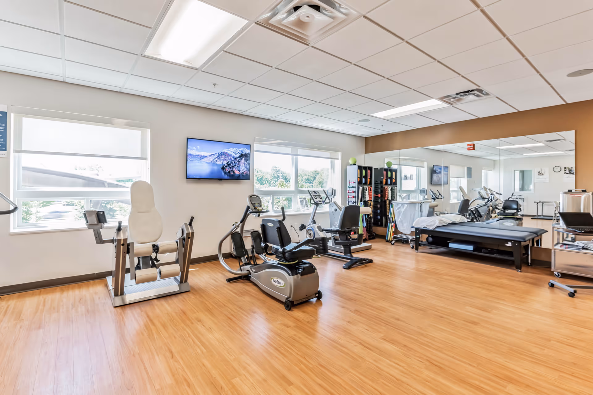 Bright rehabilitation/fitness room with exercise equipment, a treatment table, large mirror and windows.