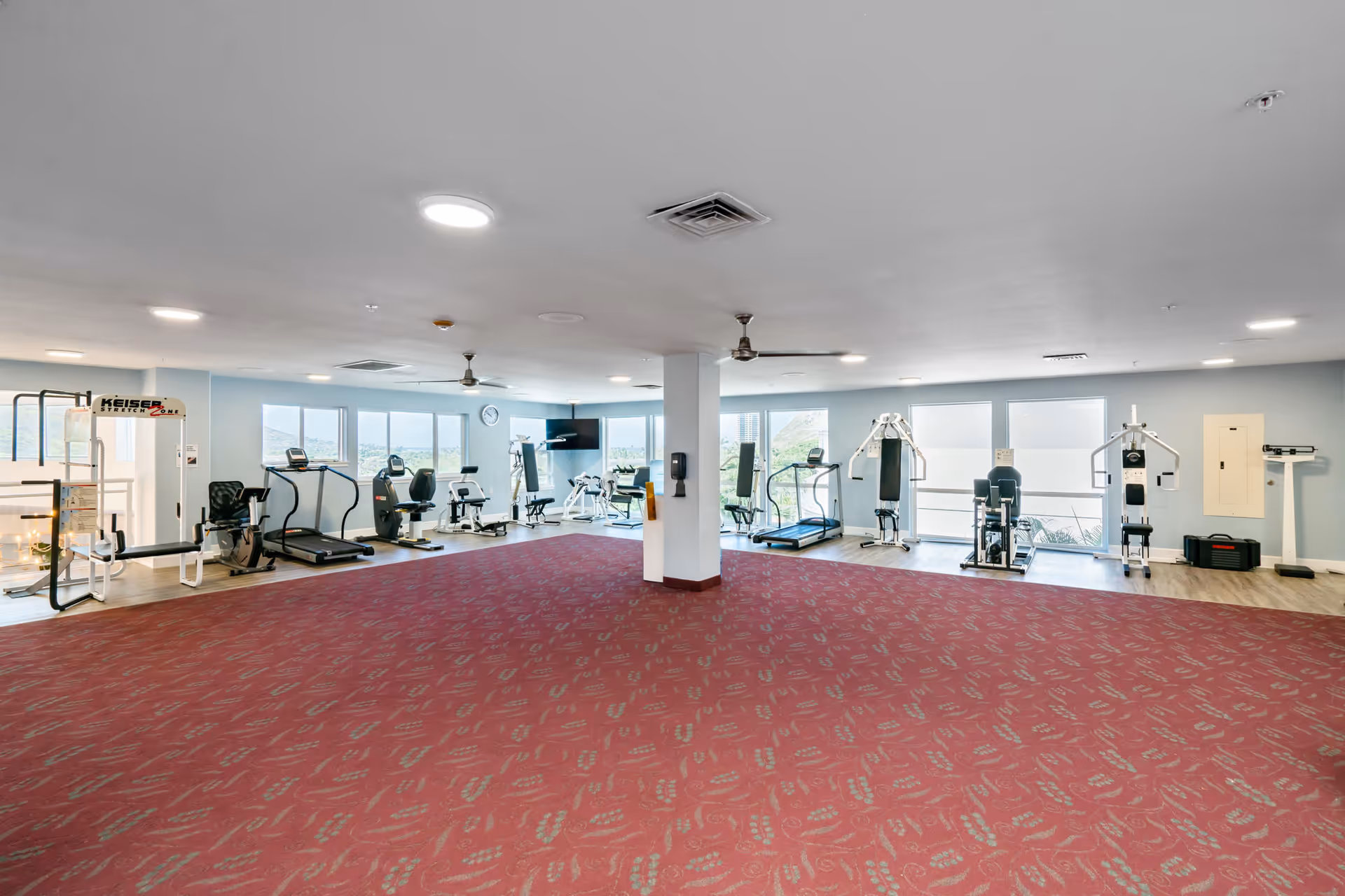 A spacious fitness room with various exercise equipment including treadmills, stationary bikes, weight machines, and a scale. The room has large windows allowing natural light and views of the outdoors. The floor is covered with red patterned carpet, and the walls are painted light blue.