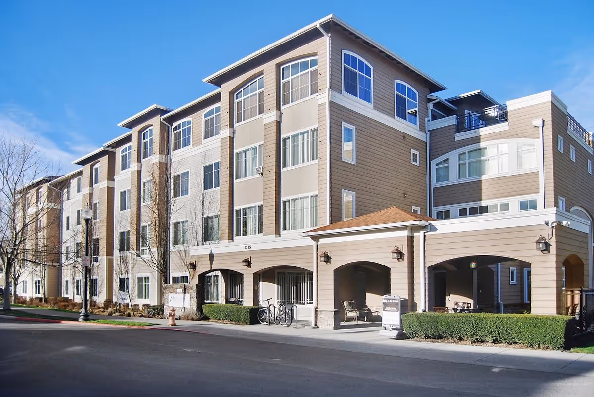 Four-story tan and white senior living building with an arched covered entrance and many windows under a clear blue sky.