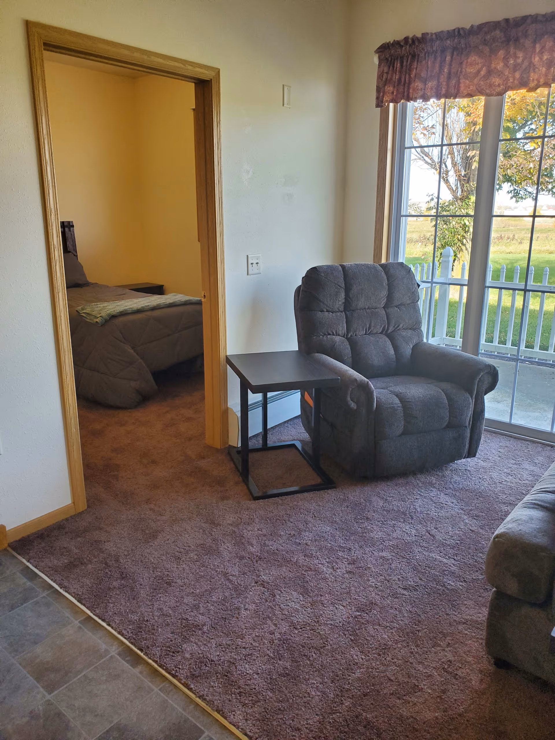 A cozy living room area with a gray recliner chair and a small black side table on a purple carpet. To the left, there is an open doorway leading to a bedroom with a bed covered in a gray comforter. On the right, there is a large window with a view of a white picket fence and green grass outside.