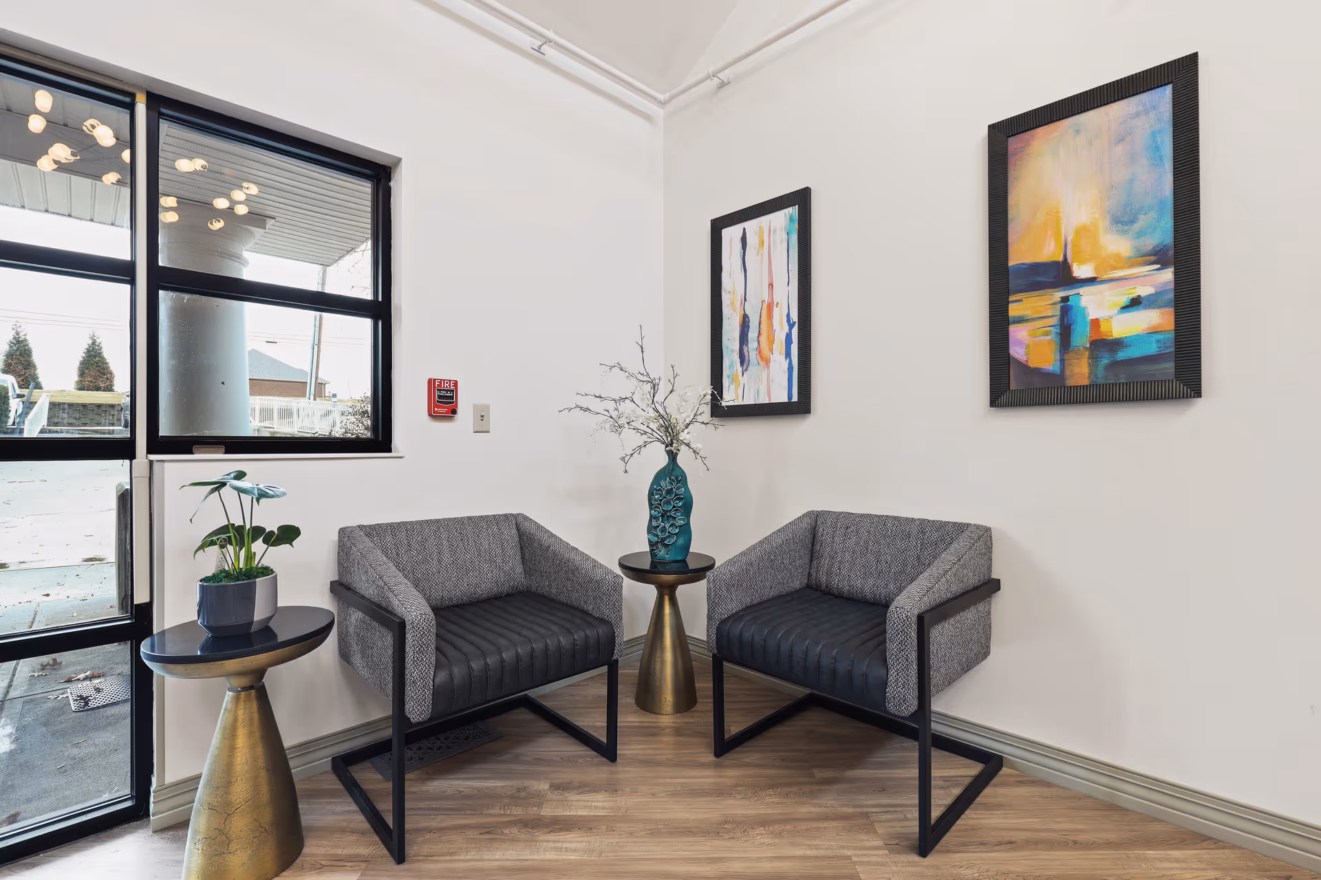 A small seating area with two modern gray armchairs with black metal frames, separated by a small round table with a blue decorative vase holding branches. Another small round table with a potted plant is next to the left chair. The room has light-colored walls with two colorful abstract paintings hanging on them, and a large window and glass door showing an outdoor view.