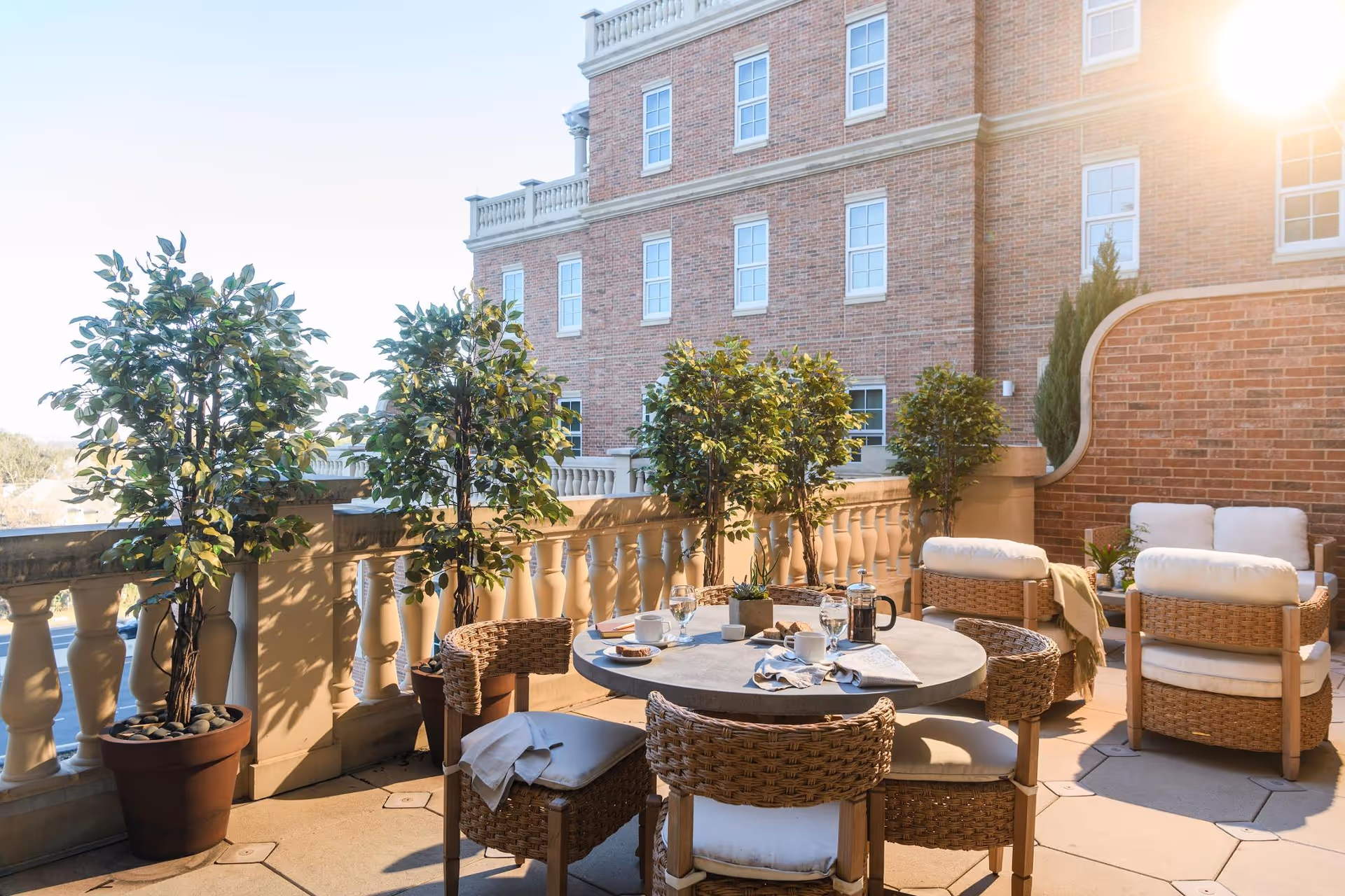 Sunny outdoor terrace with a round table, wicker chairs, potted trees, and cushioned seating against a brick building.