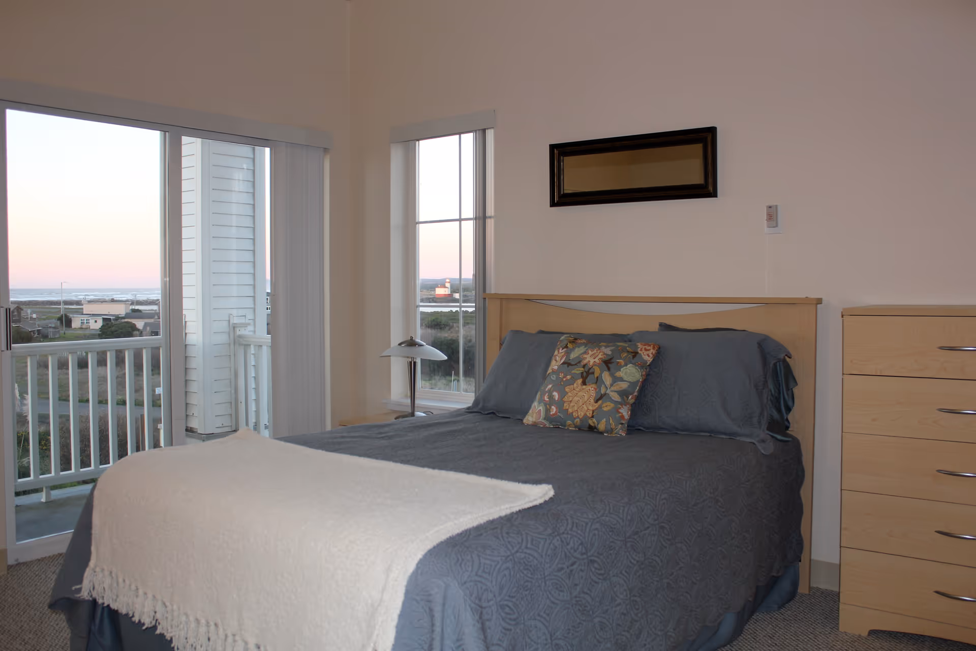A bedroom with a neatly made bed featuring a blue patterned bedspread, a white throw blanket, and a floral decorative pillow. There is a wooden headboard, a wooden dresser with silver handles, a small bedside table with a lamp, and a rectangular mirror on the wall. Large windows and a sliding glass door provide a view of an outdoor balcony and a distant lighthouse near the ocean at sunset.