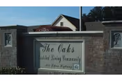 Stone entrance sign for The Oaks Assisted Living Community with a building and trees in the background under a clear sky.