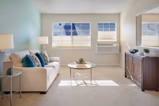 A bright living room with a white sofa adorned with blue and beige cushions, a round glass coffee table with a small plant on it, a wooden sideboard with decorative items, a large mirror above the sideboard, two windows with blinds, and a wall-mounted air conditioning unit.