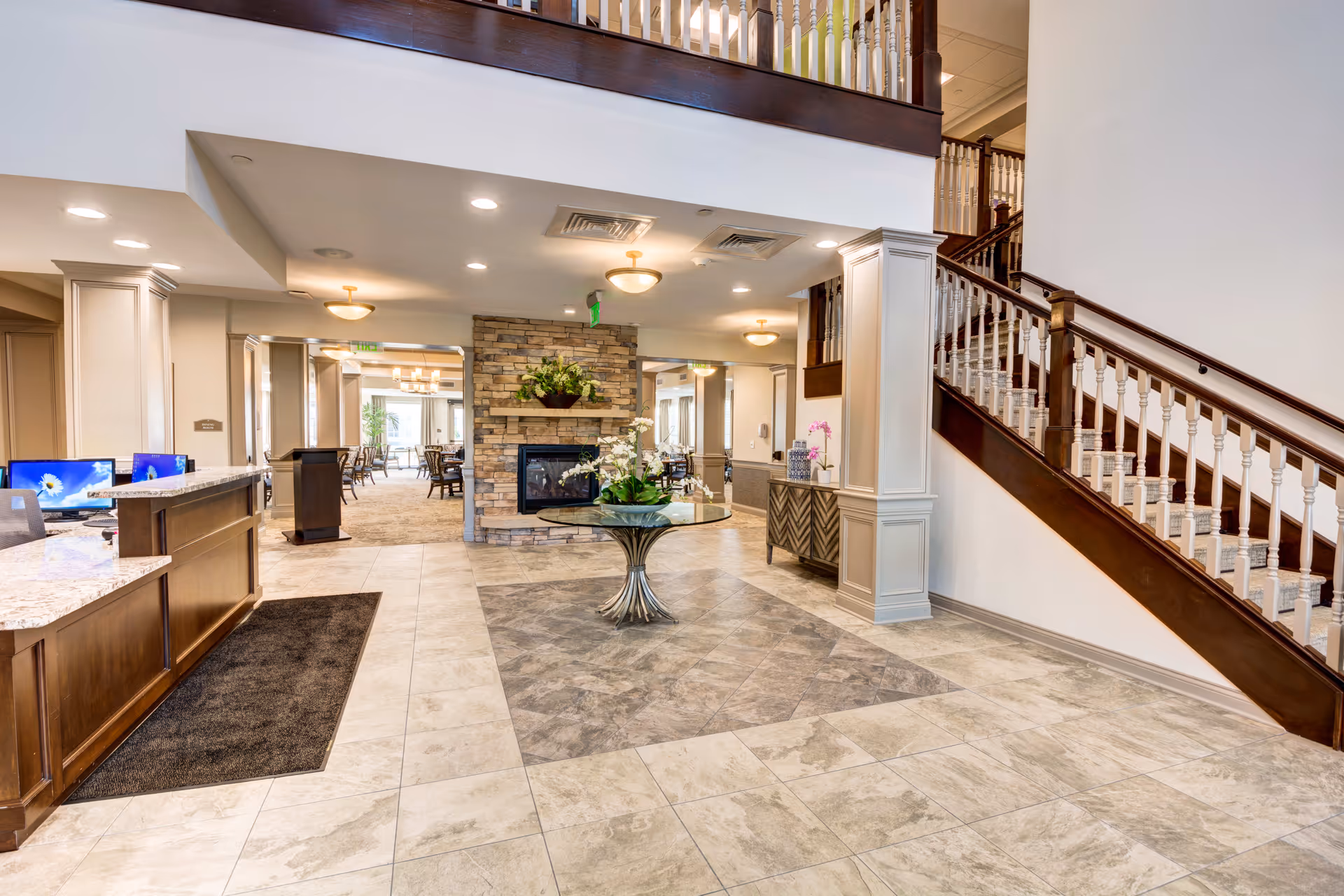 Spacious and well-lit interior lobby area of a senior living facility featuring a tiled floor with a decorative pattern in the center, a round glass table with a floral arrangement, a stone fireplace, wooden staircase with white balusters, and a reception desk with computer monitors on the left side.