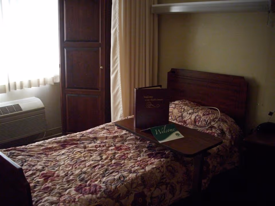 A small bedroom with a single bed covered in a floral patterned quilt. A wooden headboard is visible, and a wooden cabinet stands near a window with sheer curtains. A portable overbed table is placed on the bed, holding a welcome booklet and a green welcome card. An air conditioning unit is installed below the window.