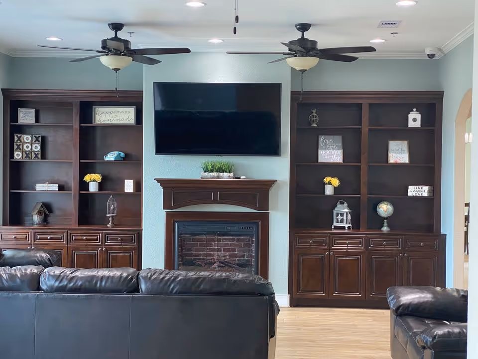 A cozy living room area with dark leather sofas facing a wall-mounted flat screen TV above a wooden fireplace. On either side of the fireplace are built-in dark wood shelves and cabinets decorated with small plants, framed quotes, a globe, and other decorative items. The ceiling has two ceiling fans with lights, and the walls are painted a soft blue color.