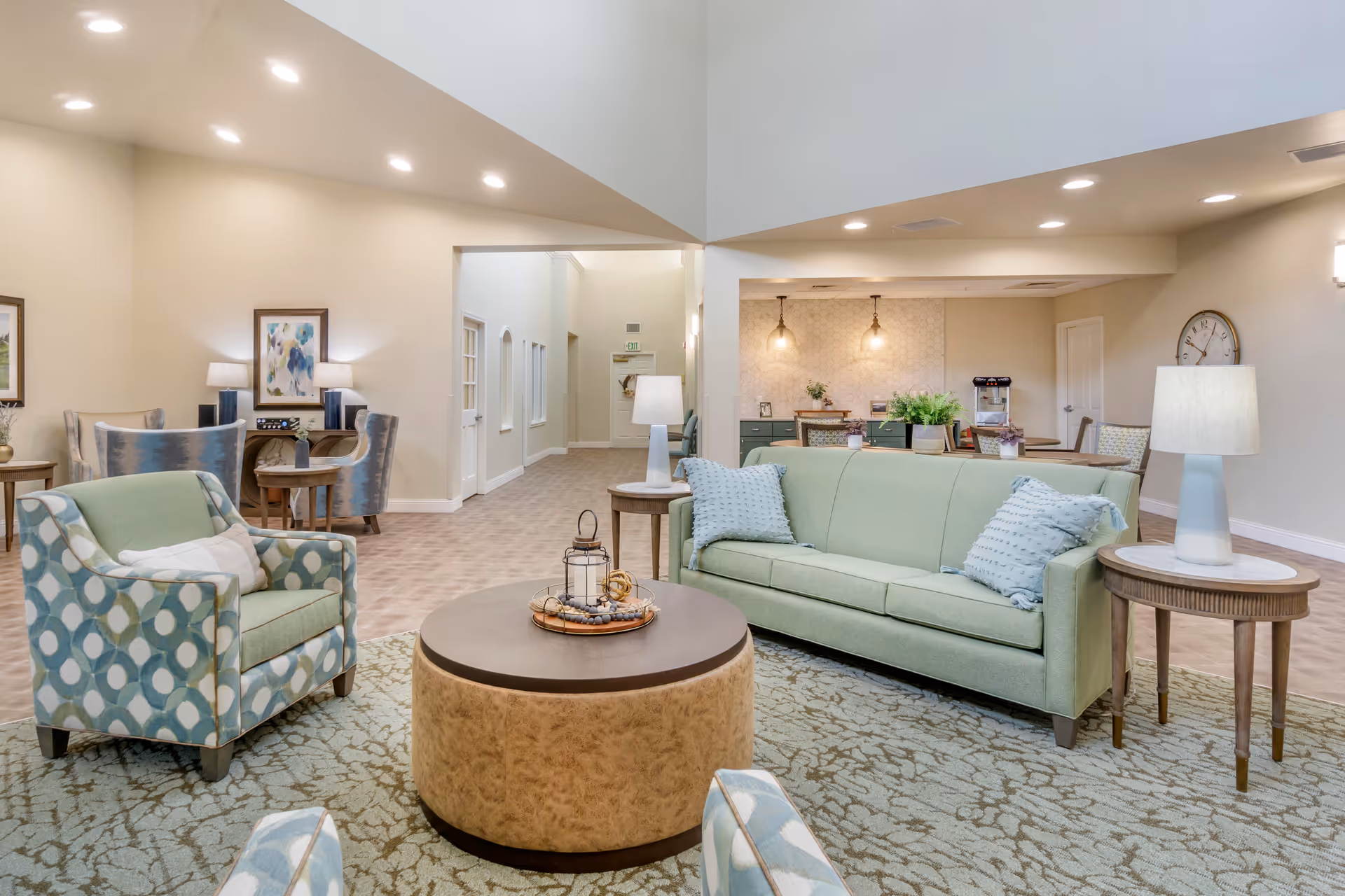 A bright and spacious living room area in a senior living facility featuring a light green sofa with two blue pillows, a patterned armchair, a round coffee table with decorative items, side tables with lamps, and additional seating and tables in the background. The room has beige walls, recessed lighting, and a patterned carpet.
