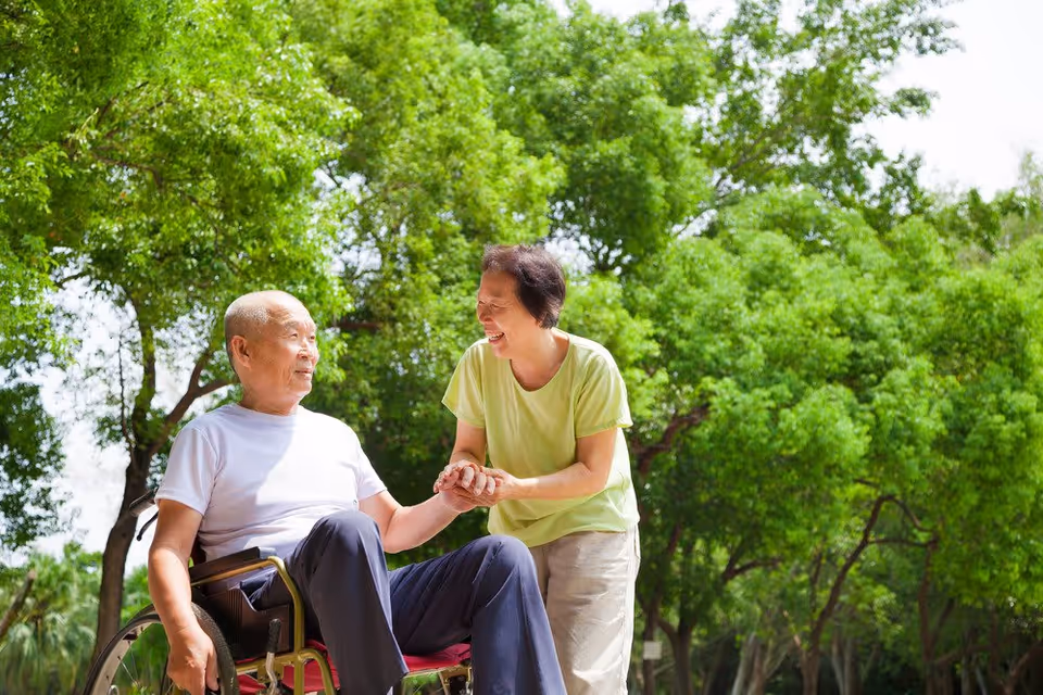 An elderly man in a wheelchair holding hands and smiling with an elderly woman standing beside him in a lush green outdoor park setting with trees in the background.