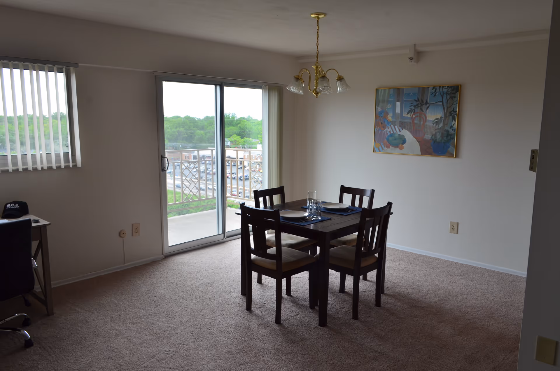 A dining area with a dark wooden table set for four with plates and glasses. The room has beige carpet, white walls, a hanging light fixture, and a framed painting on the wall. There is a sliding glass door leading to a balcony with a view of trees and buildings outside. A window with vertical blinds is on the left side, and a small desk with a chair is partially visible.