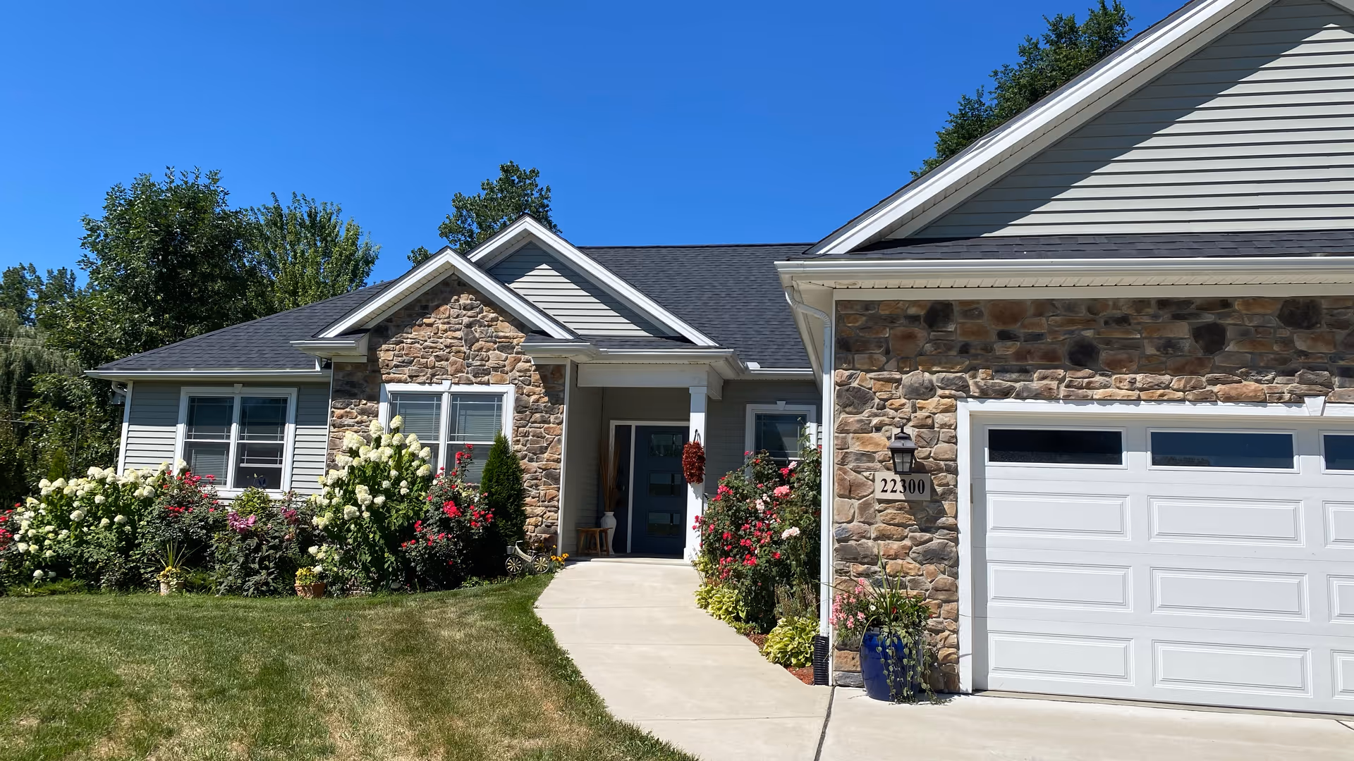 Front exterior of a single-story stone-and-siding house with a garage, driveway, and landscaped flowerbeds under a clear blue sky.