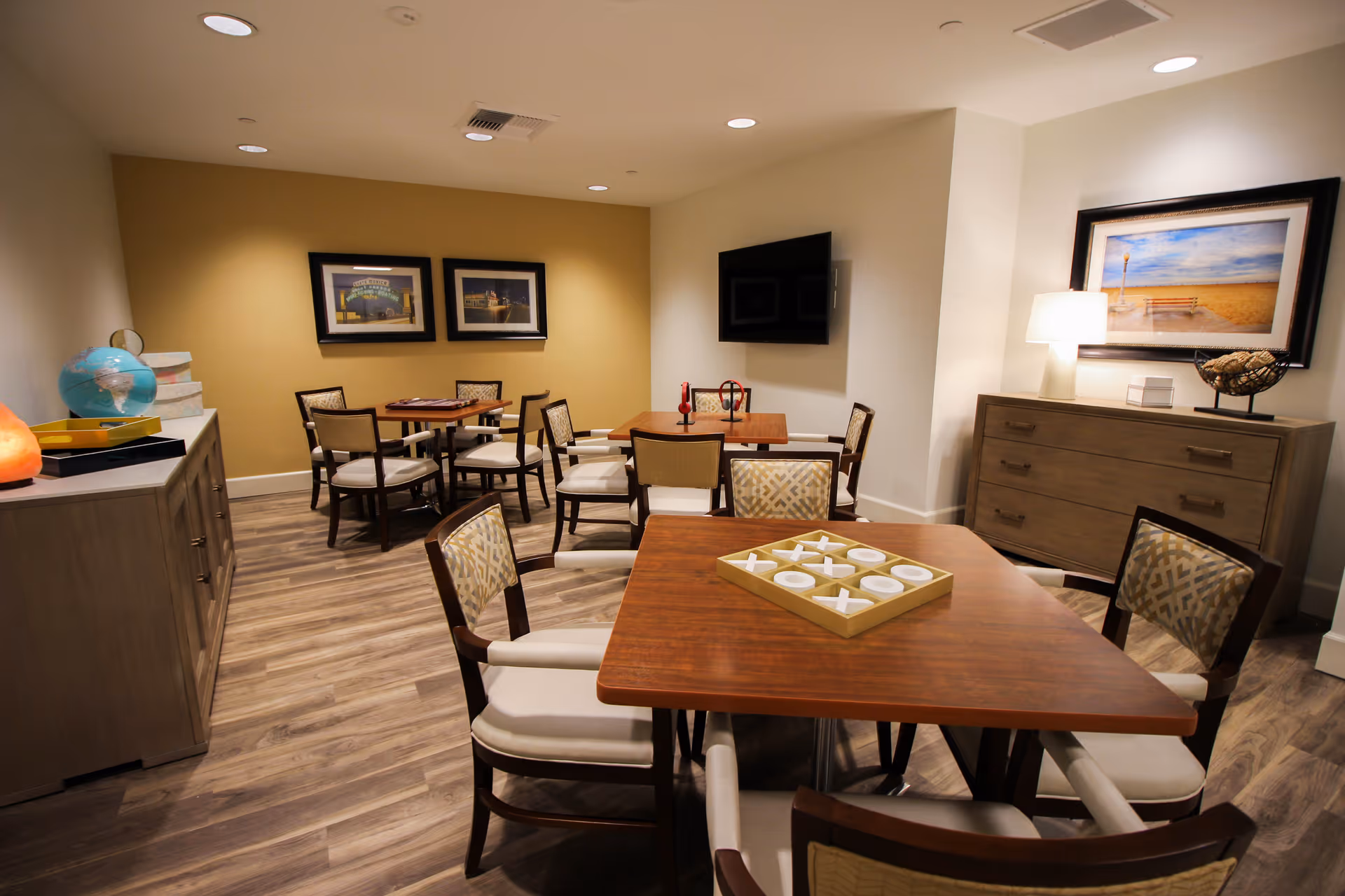 A cozy common area with several wooden tables and cushioned chairs arranged for socializing or games. One table in the foreground has a tic-tac-toe game set on it. The room features wood flooring, warm lighting, framed pictures on the walls, a flat-screen TV mounted on the wall, and a sideboard with a lamp and decorative items.