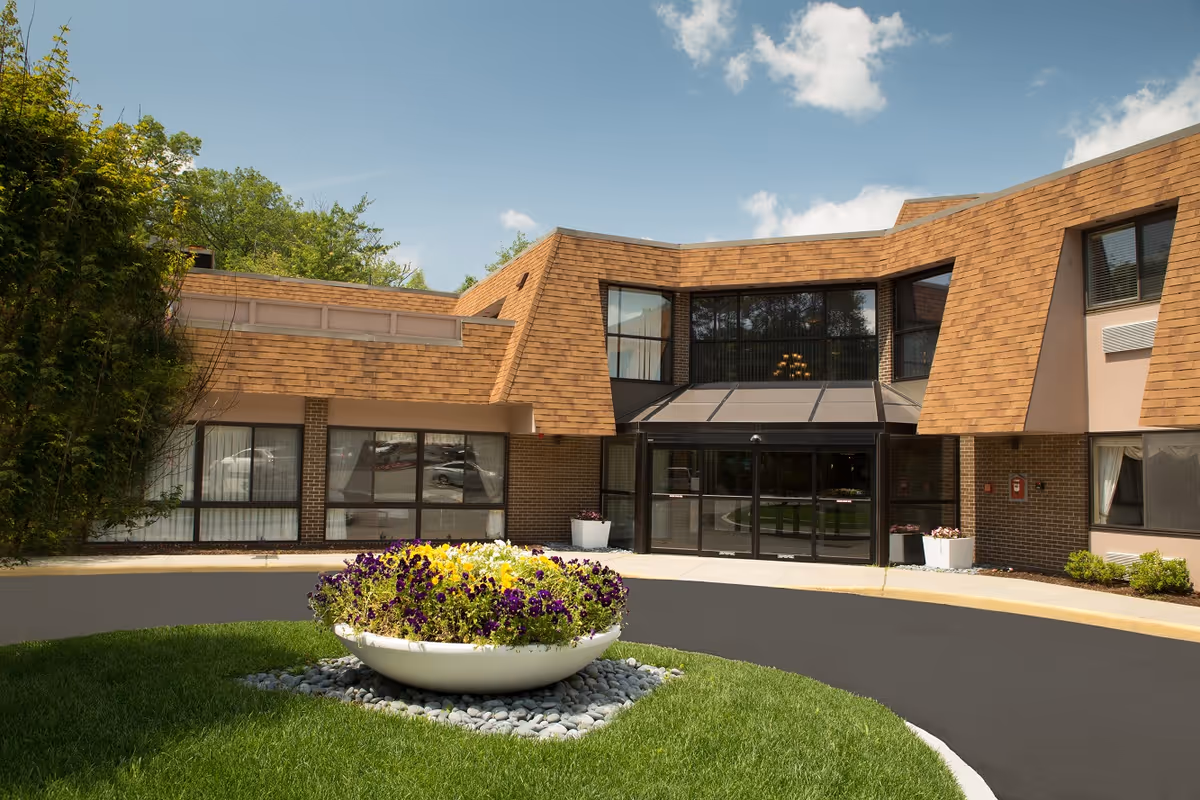Exterior view of Bear Mountain at Reading facility showing the entrance with large glass doors, brown shingled roof, brick walls, and a circular driveway with a flower planter in the center. Trees and blue sky with clouds are visible in the background.