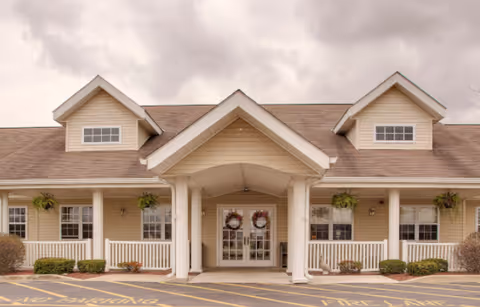 Front entrance of a single-story beige senior living facility with a covered porch, white columns, double glass doors with wreaths, and dormer windows.