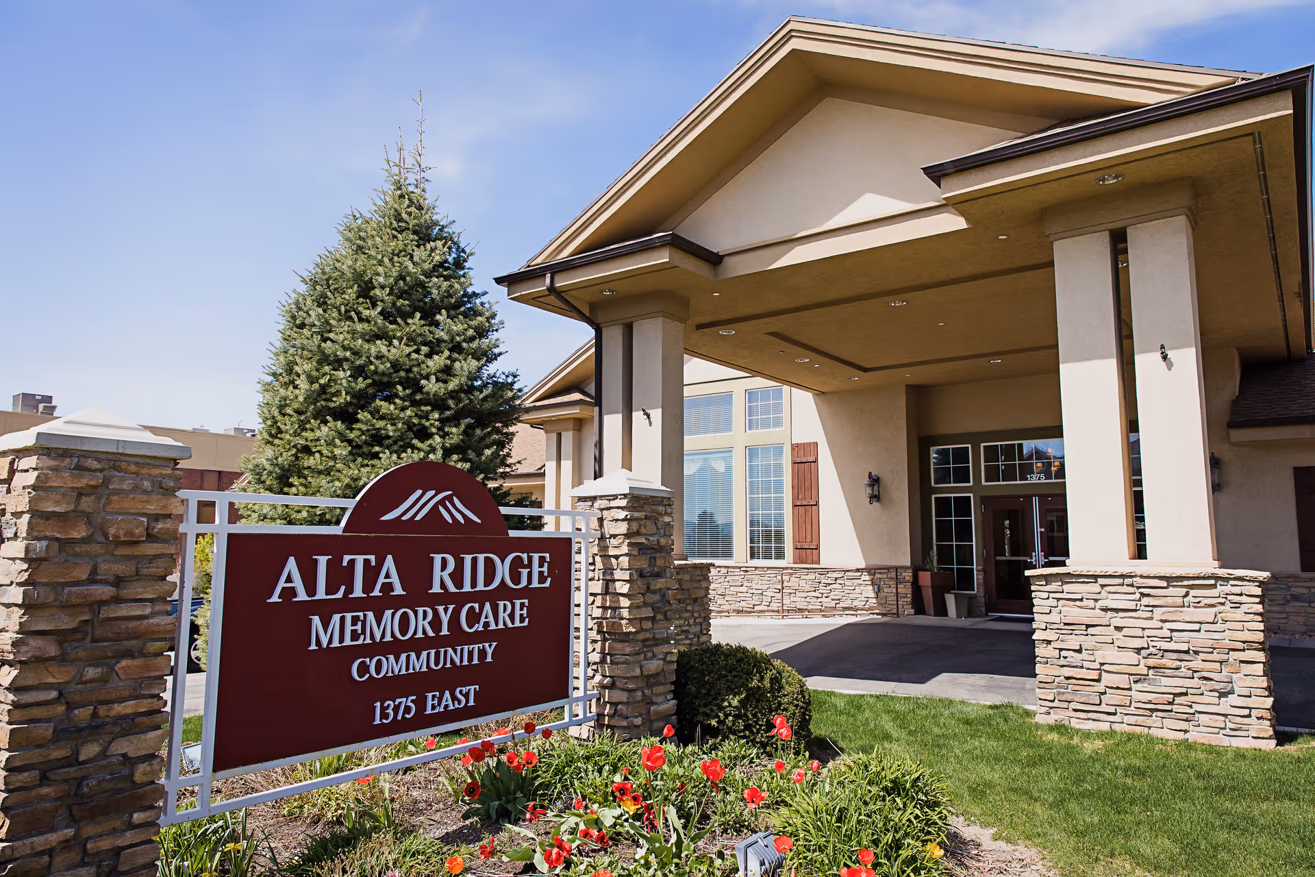 Entrance of Alta Ridge Memory Care Community building with a covered driveway, stone pillars, and a landscaped garden with red flowers and green bushes. A large sign in front reads 'Alta Ridge Memory Care Community 1375 East'.