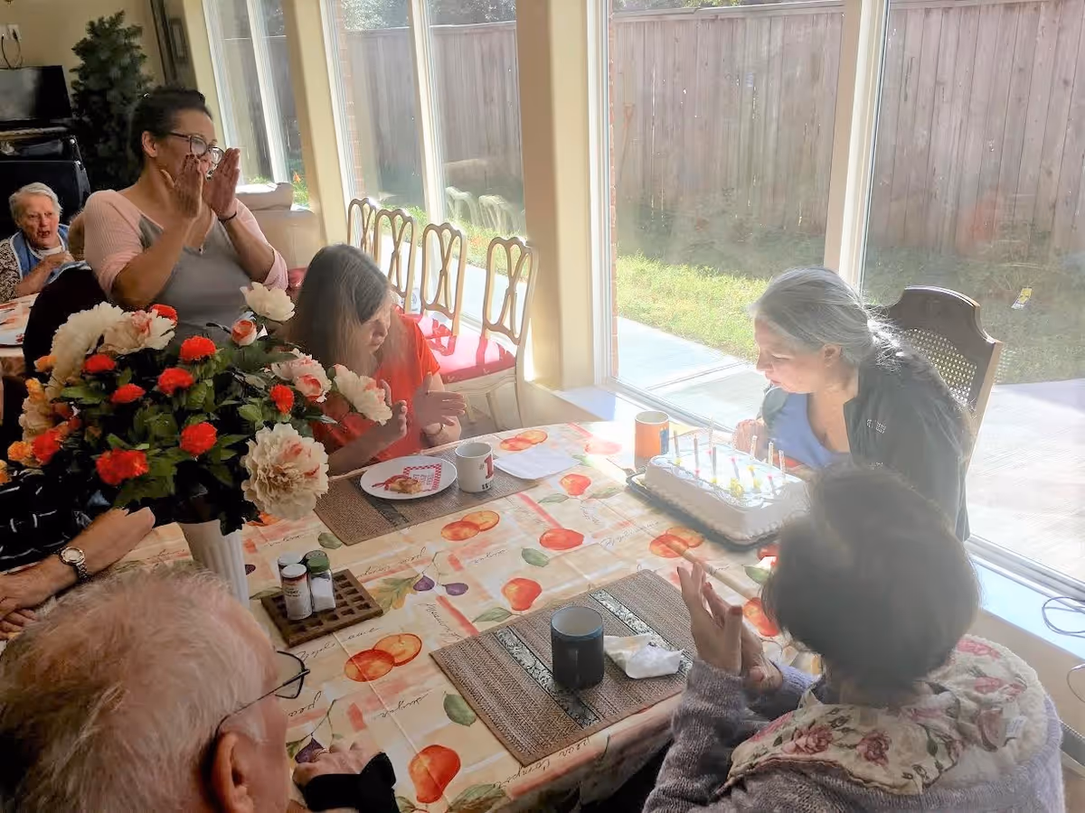 A group of elderly people and a caregiver gathered around a dining table with a birthday cake. One elderly woman is blowing out candles on the cake while others watch and clap. The room has large windows letting in natural light and a vase of flowers on the table.