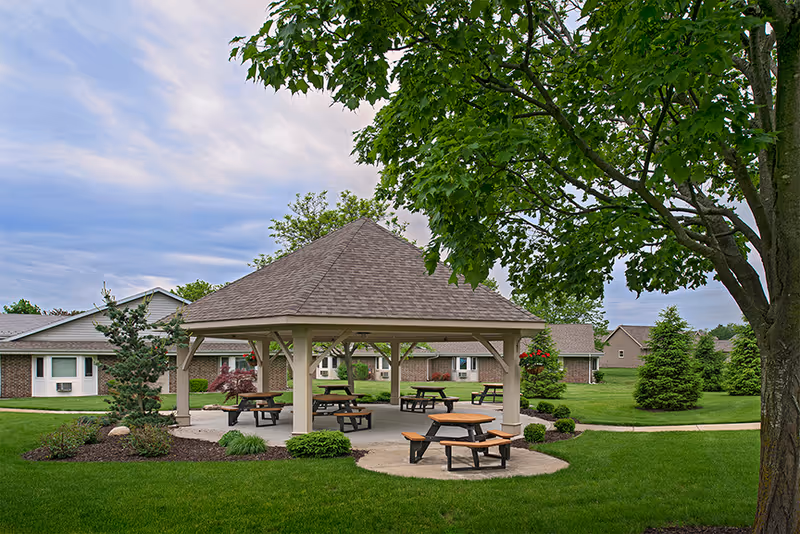 Outdoor pavilion with picnic tables and benches surrounded by green grass, trees, and shrubs at American House Holland senior living facility under a partly cloudy sky.