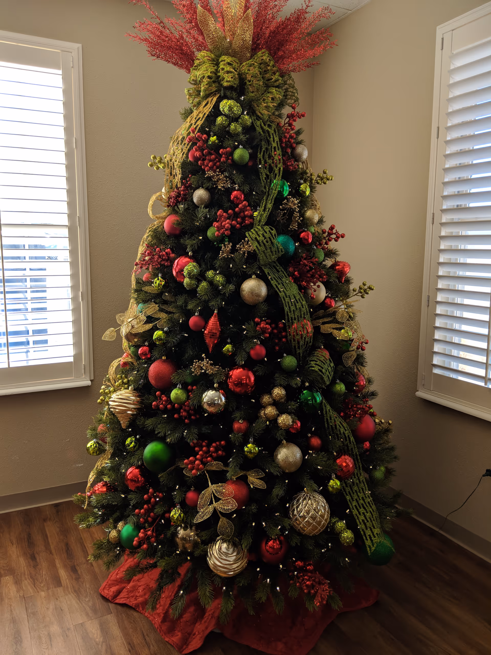 A decorated Christmas tree with red, green, and gold ornaments, ribbons, and berries, standing on a wooden floor between two windows with white blinds in a room with beige walls.