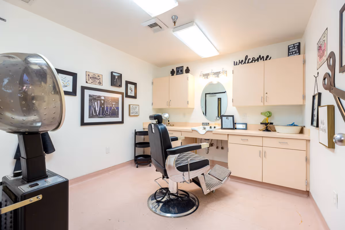 Interior view of a small barber or hair salon room with a black barber chair in the center, a hair dryer hood on the left, beige cabinets and countertop with a sink, an oval mirror, and various framed pictures and decorations on the walls including a large pair of scissors and a welcome sign.