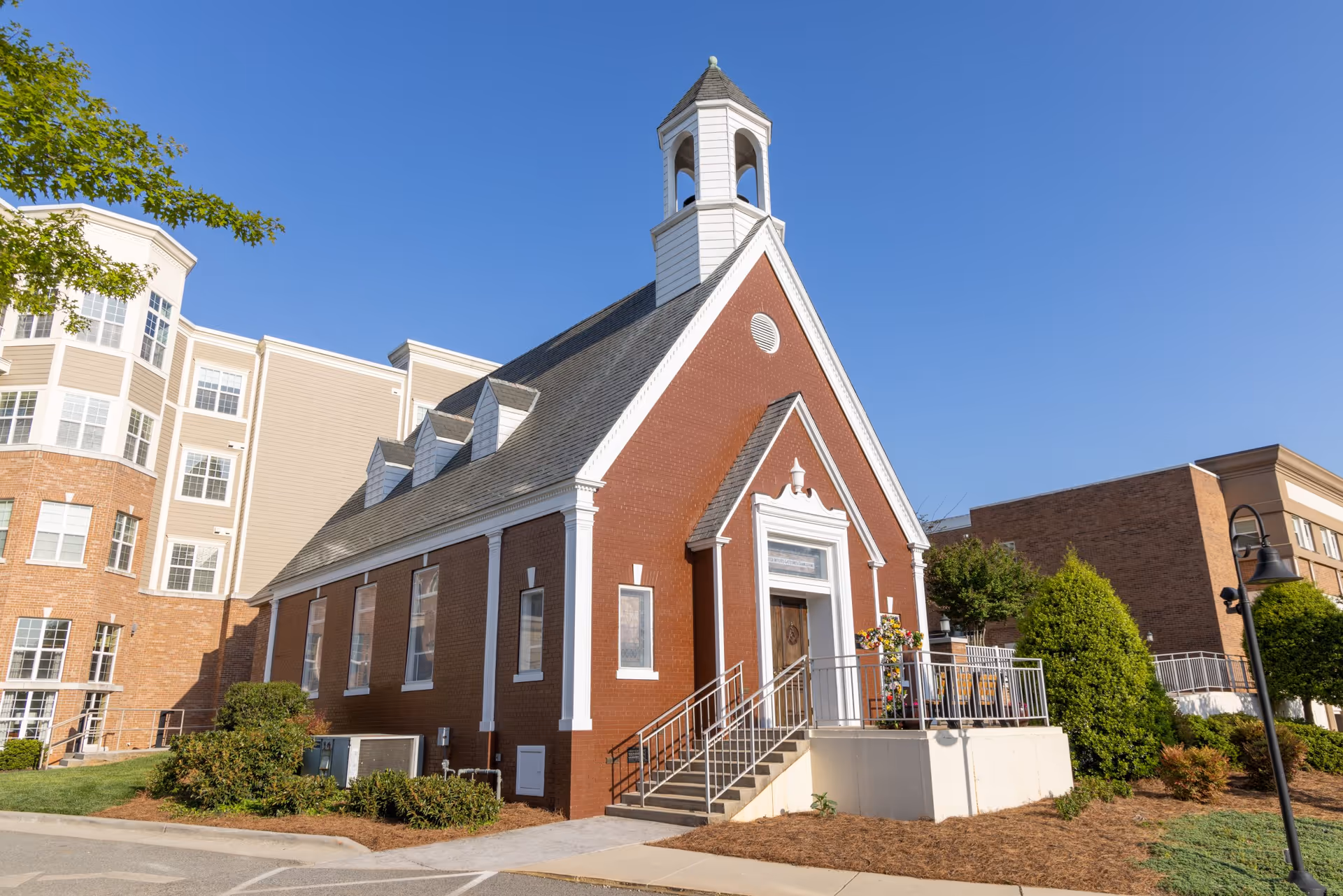 A small red-brick chapel-style building with a white steeple and entrance steps beside a multi-story residential building under a clear blue sky.
