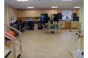 Rehabilitation/activity room with parallel bars and exercise equipment, a table with chairs, computers and cabinets along the back wall.