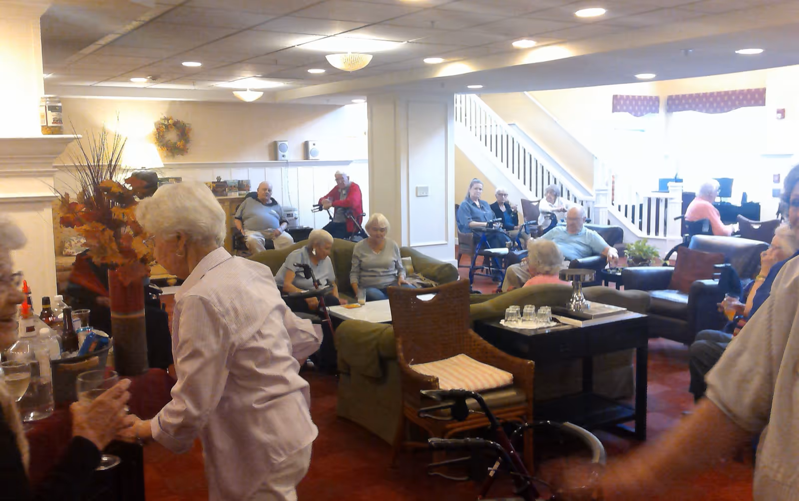 Elderly residents socializing in a senior living facility lounge with chairs, tables, and walkers.