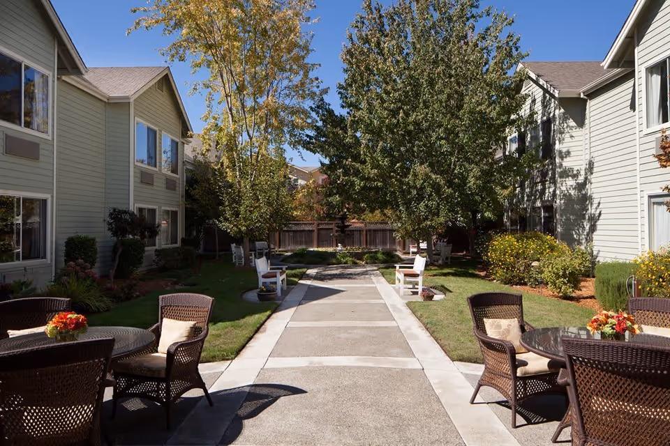 Outdoor courtyard area at Windsong of Sonoma Senior Living with a paved walkway lined by trees and bushes. There are white benches along the path and round tables with wicker chairs and floral centerpieces on either side. The courtyard is surrounded by two-story beige buildings with multiple windows under a clear blue sky.