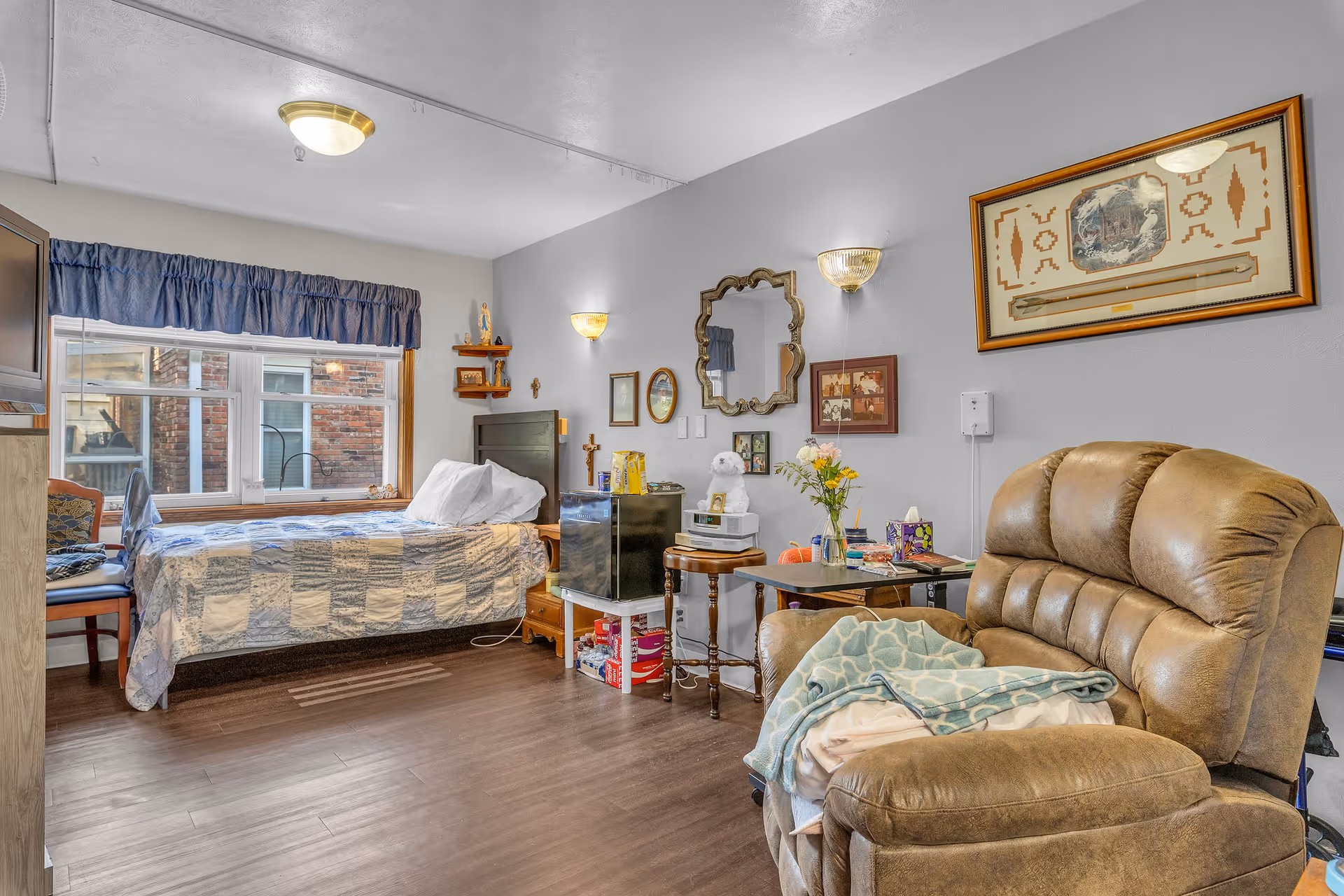 A cozy room with a single bed covered in a patchwork quilt, a large window with blue valance, a brown recliner chair with a folded blanket on it, and various framed pictures and a mirror on the light gray walls. There is a small table with flowers, tissues, and other personal items, and a mini fridge next to the bed.