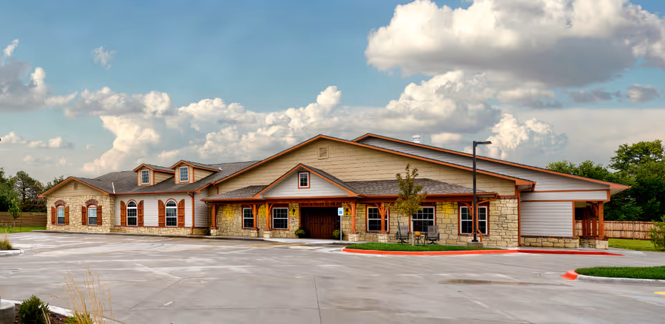 Exterior view of a single-story assisted living facility building with stone and wood siding, multiple windows, a covered entrance, and a large paved parking area under a partly cloudy sky.