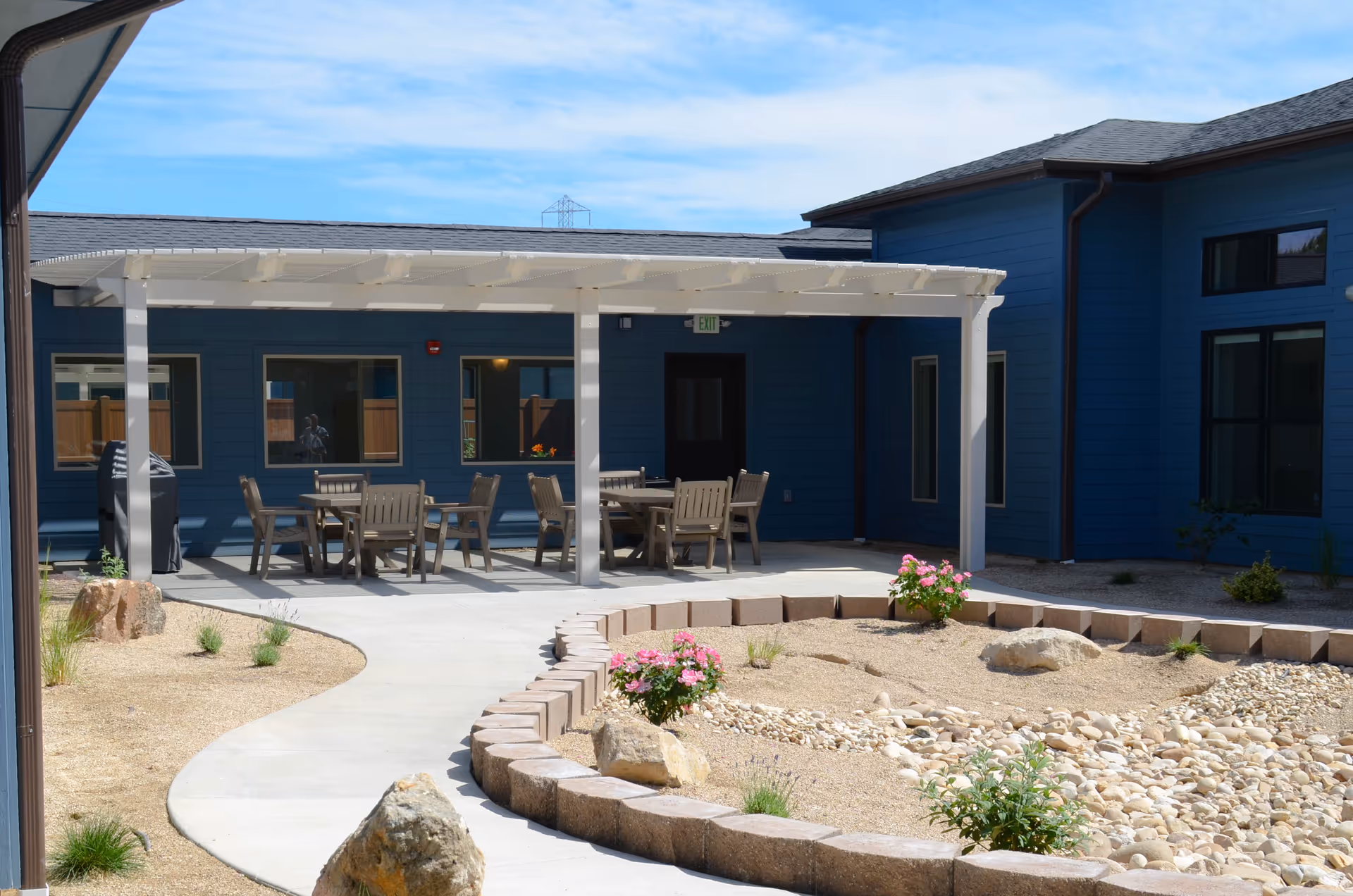 Outdoor patio area at Aspen Valley Senior Living with a white pergola covering several wooden tables and chairs. The patio is surrounded by a landscaped garden with rocks, small plants, and flowers. The building exterior is painted blue with multiple windows and a door under the pergola.