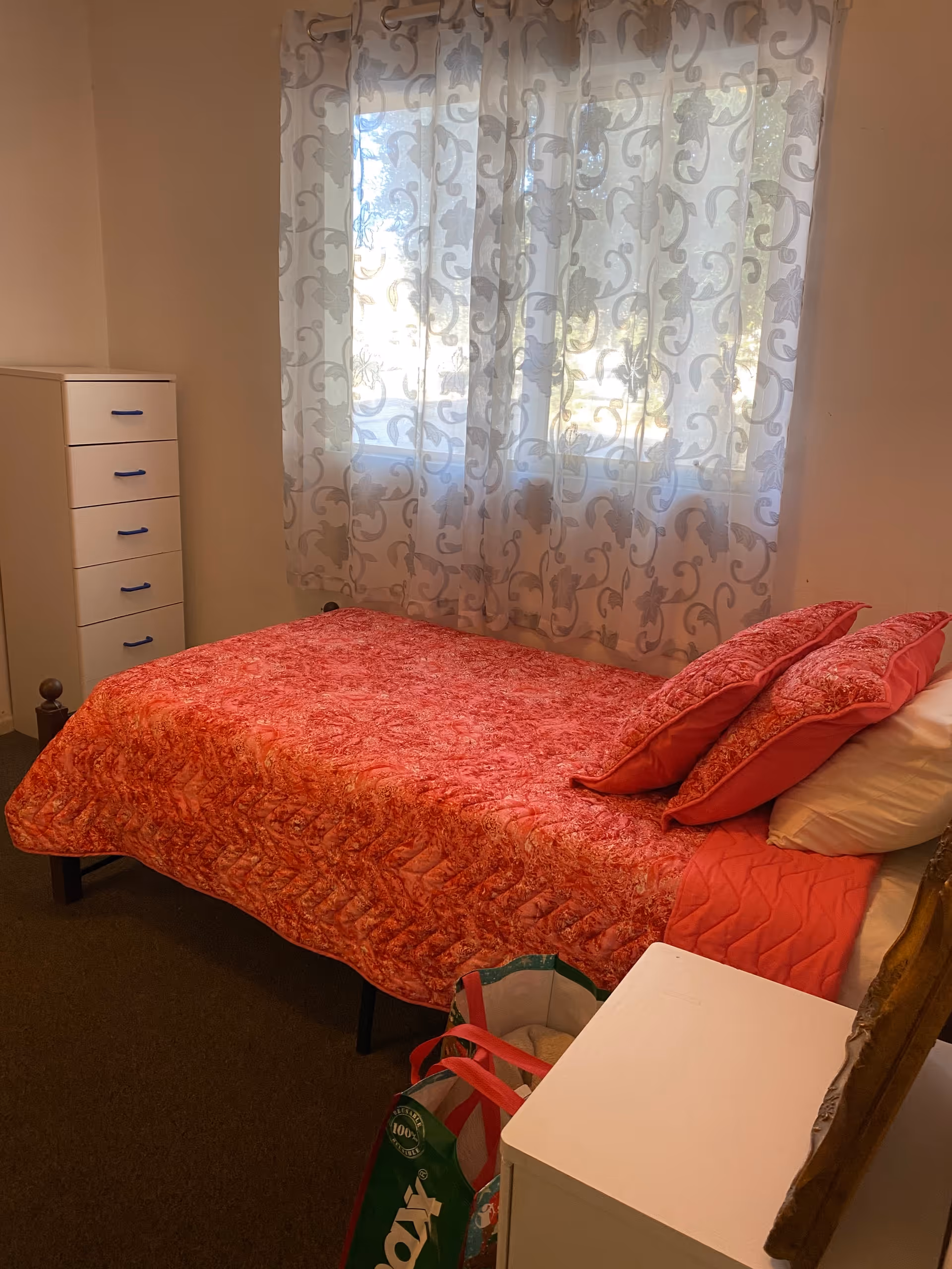 A small bedroom with a single bed covered in a red patterned quilt and matching pillows. The bed is positioned next to a window with sheer white curtains featuring a floral design. There is a white chest of drawers with blue handles in the corner and a white nightstand beside the bed. A reusable shopping bag is placed on the floor near the nightstand.