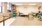 A physical therapy or rehabilitation room with parallel bars for walking exercises, a treatment table, chairs, exercise balls, and various therapy equipment along the walls. The room is well-lit with large windows and overhead lights.