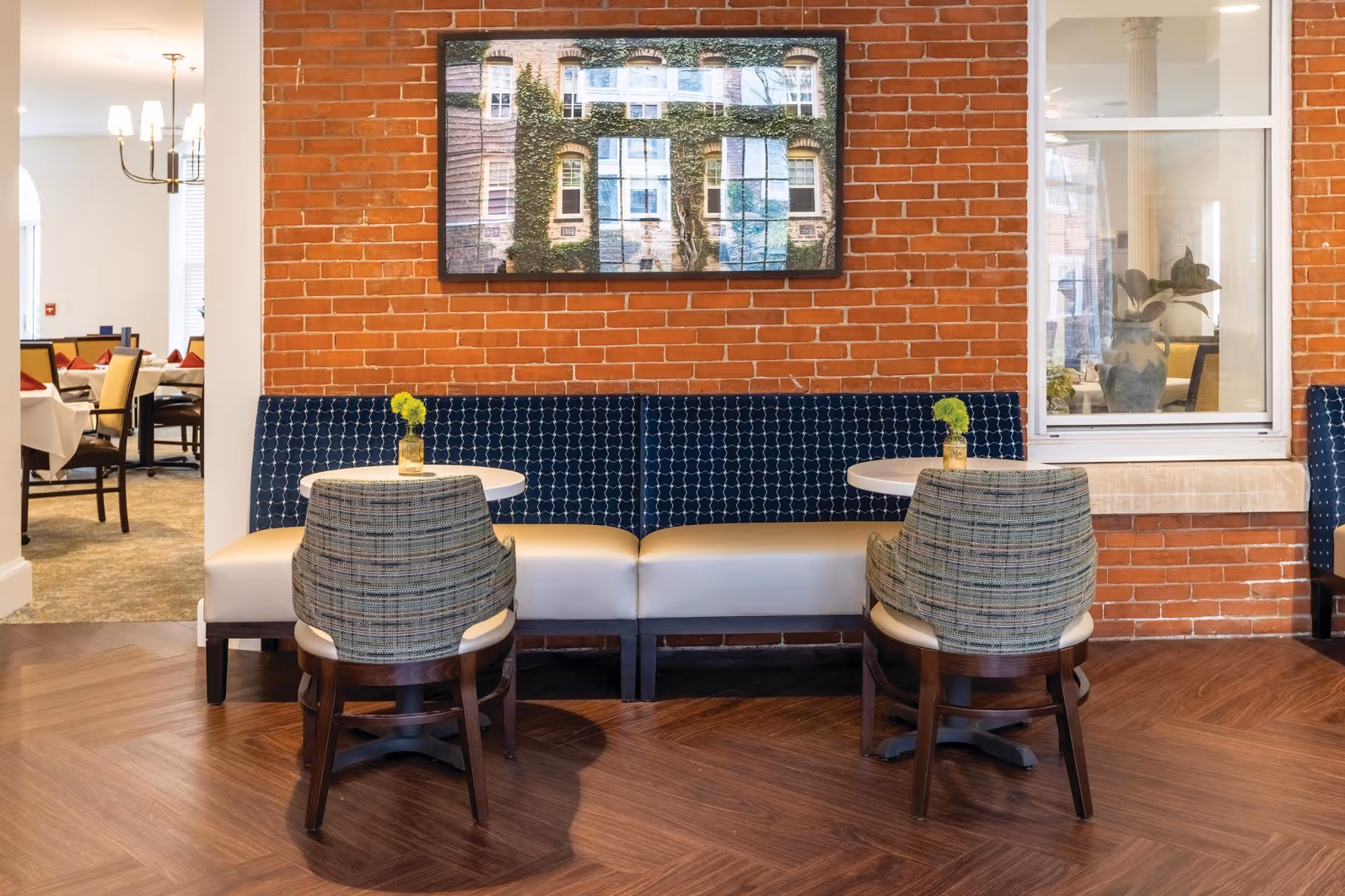 A cozy seating area with two small round tables, each accompanied by a cushioned bench and a patterned chair. The backdrop features an exposed red brick wall with a framed picture of a building covered in ivy. To the left, a dining area with tables and chairs is visible, illuminated by a chandelier.
