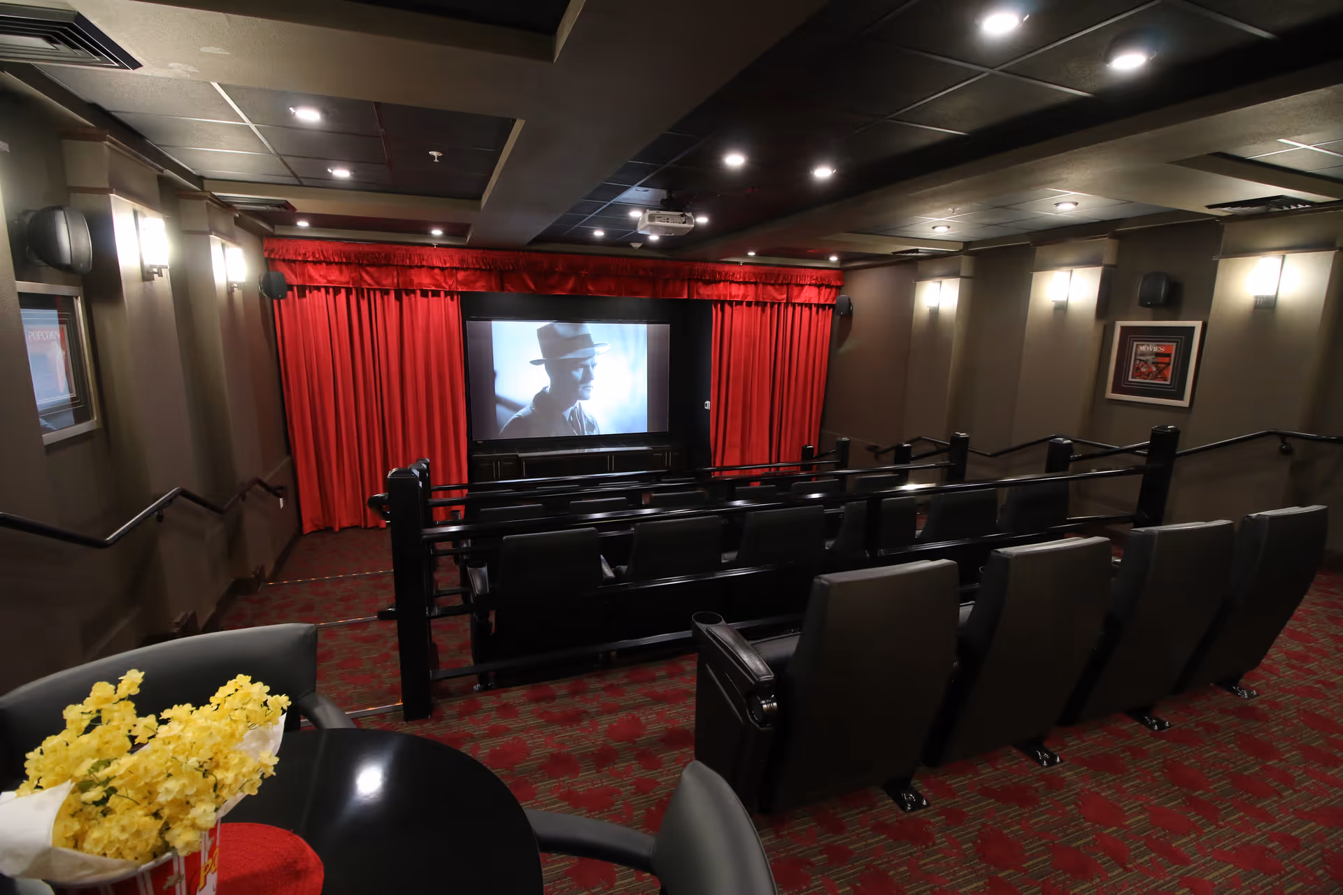 Interior view of a small movie theater room with rows of black cushioned seats facing a screen showing a black and white image of a man wearing a hat. The room has red curtains around the screen, dim wall lights, and a patterned red carpet. In the foreground, there is a round table with a bucket of yellow flowers and some chairs.