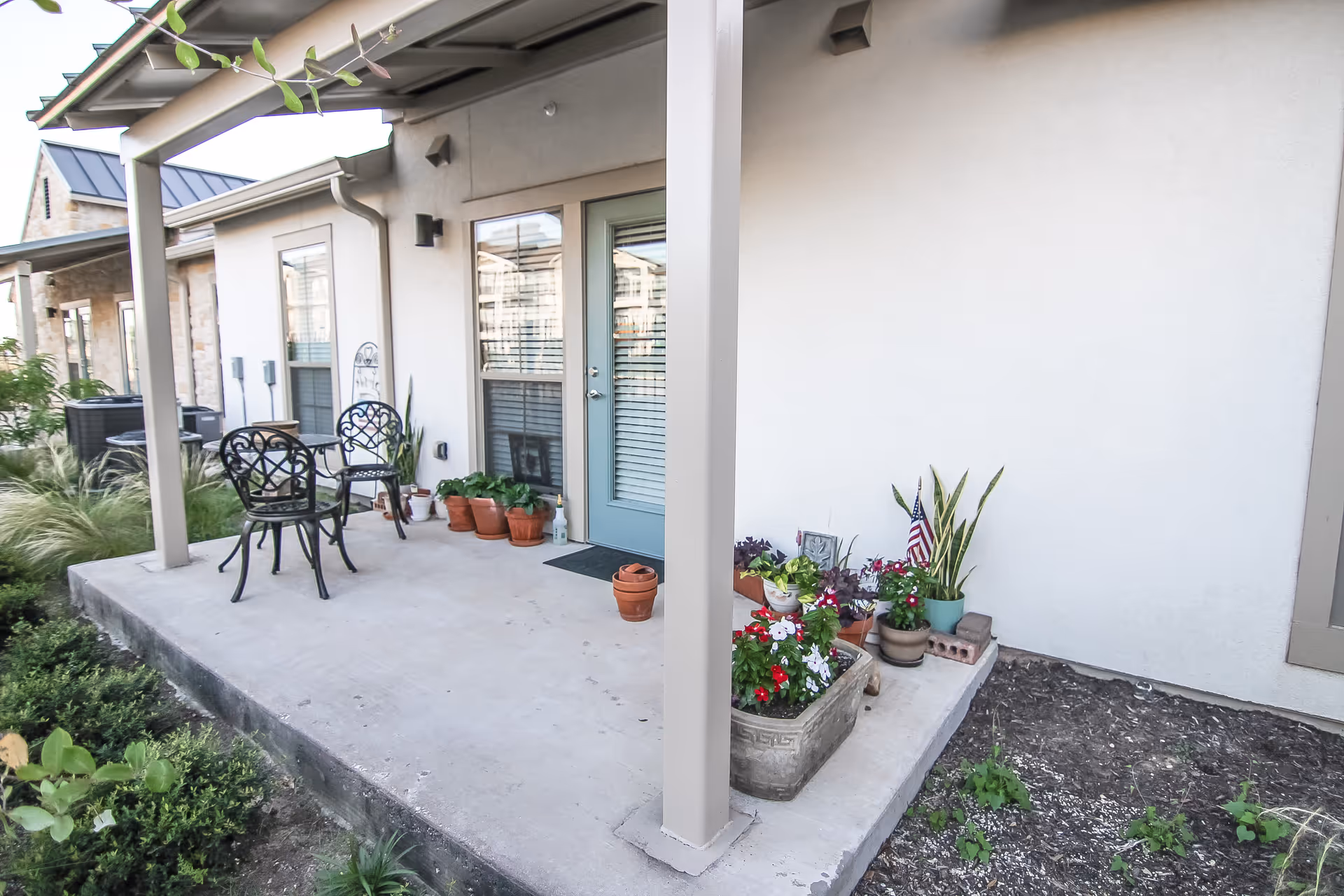 A covered patio area outside a residential unit with two black metal chairs and a small table. Several potted plants and flowers are arranged near the light blue door with glass panels. The patio is surrounded by greenery and garden beds.