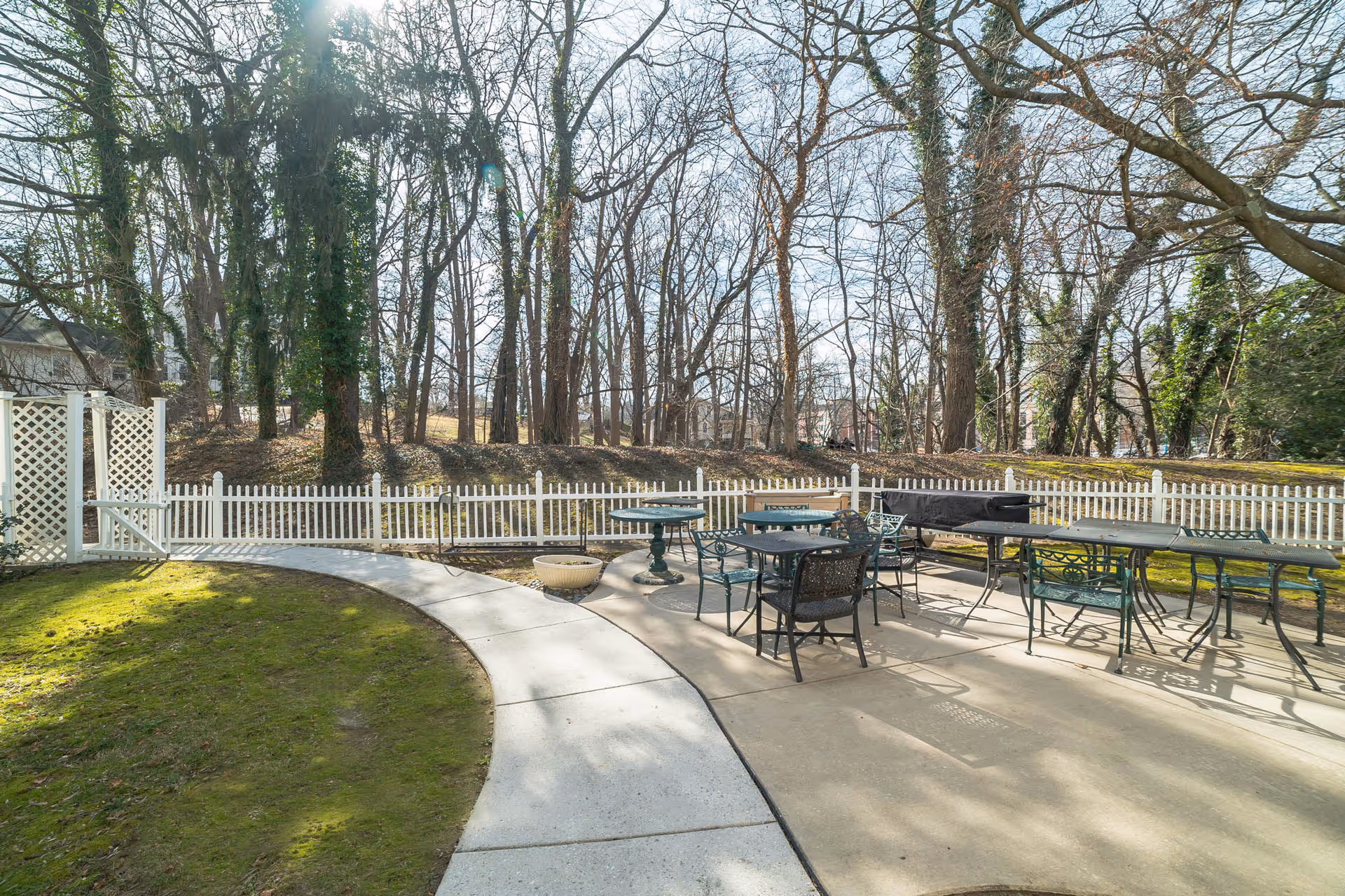 Outdoor patio area with several metal tables and chairs on a concrete surface, surrounded by a white picket fence. There is a curved concrete walkway leading to the patio, and leafless trees are visible in the background under a clear sky.