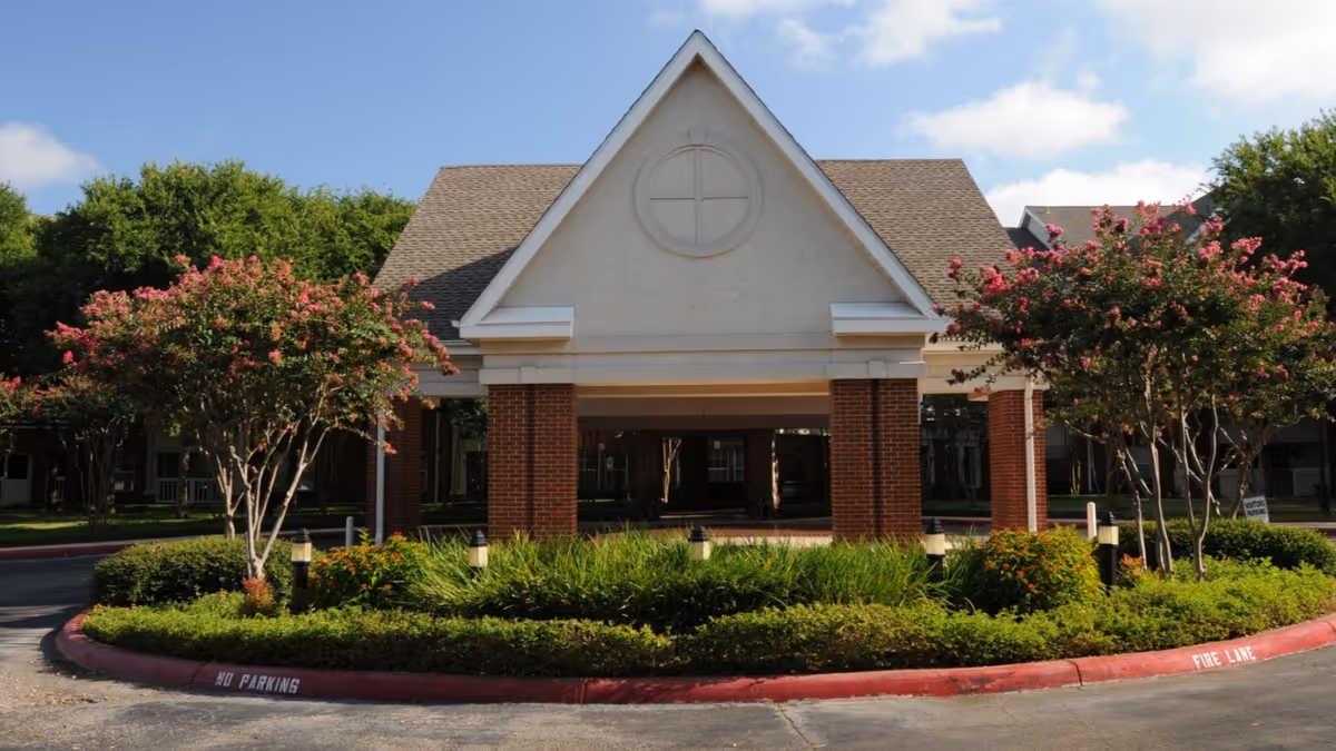 Front entrance of a building with a peaked roof supported by brick columns, surrounded by landscaped greenery and flowering trees under a blue sky with some clouds.