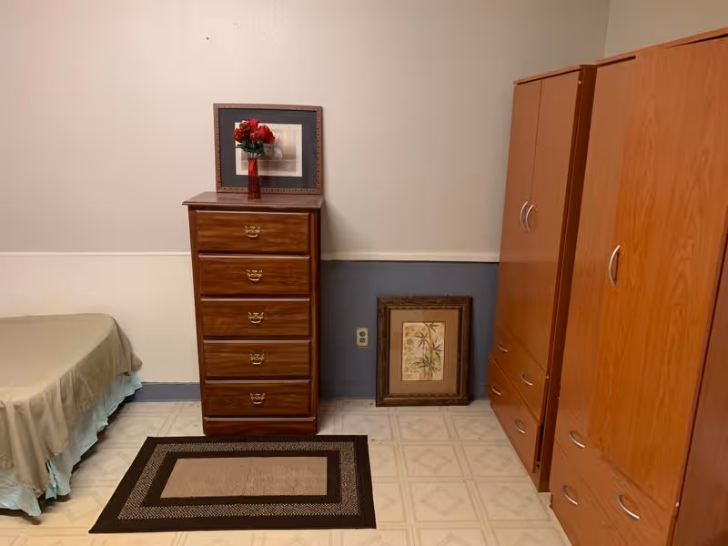A simple bedroom with a single bed covered with a beige bedspread and a light blue bed skirt. Next to the bed is a wooden chest of drawers with five drawers, on top of which is a framed picture and a red vase with red flowers. On the floor in front of the chest is a rectangular rug with a geometric pattern. To the right are two wooden wardrobes with silver handles. A framed botanical print rests against the wall between the chest and the wardrobes. The walls are painted beige on the upper half and blue on the lower half, and the floor has a light-colored tile pattern.