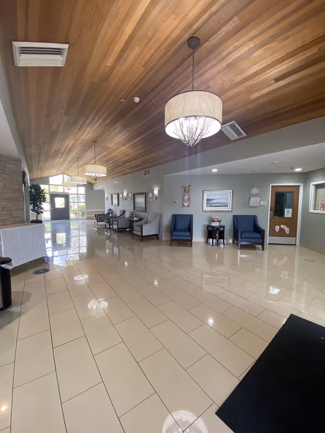 Spacious senior living facility lobby with a wood-paneled ceiling, hanging chandeliers, tiled floor, and seating along the walls.