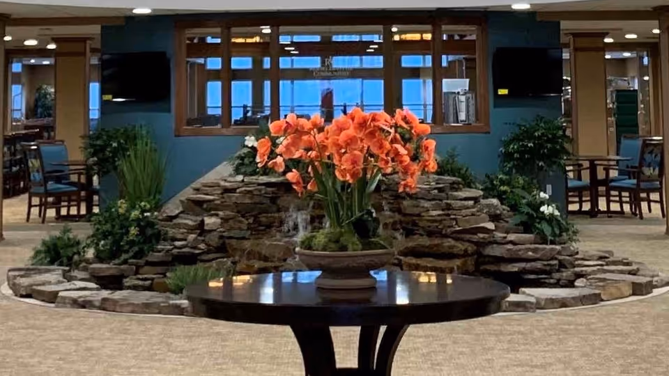 Indoor common area with a round table in the foreground holding a pot of vibrant orange flowers. Behind the table is a decorative rock water feature with greenery. The background shows seating areas with chairs and tables, two wall-mounted televisions, and large windows.
