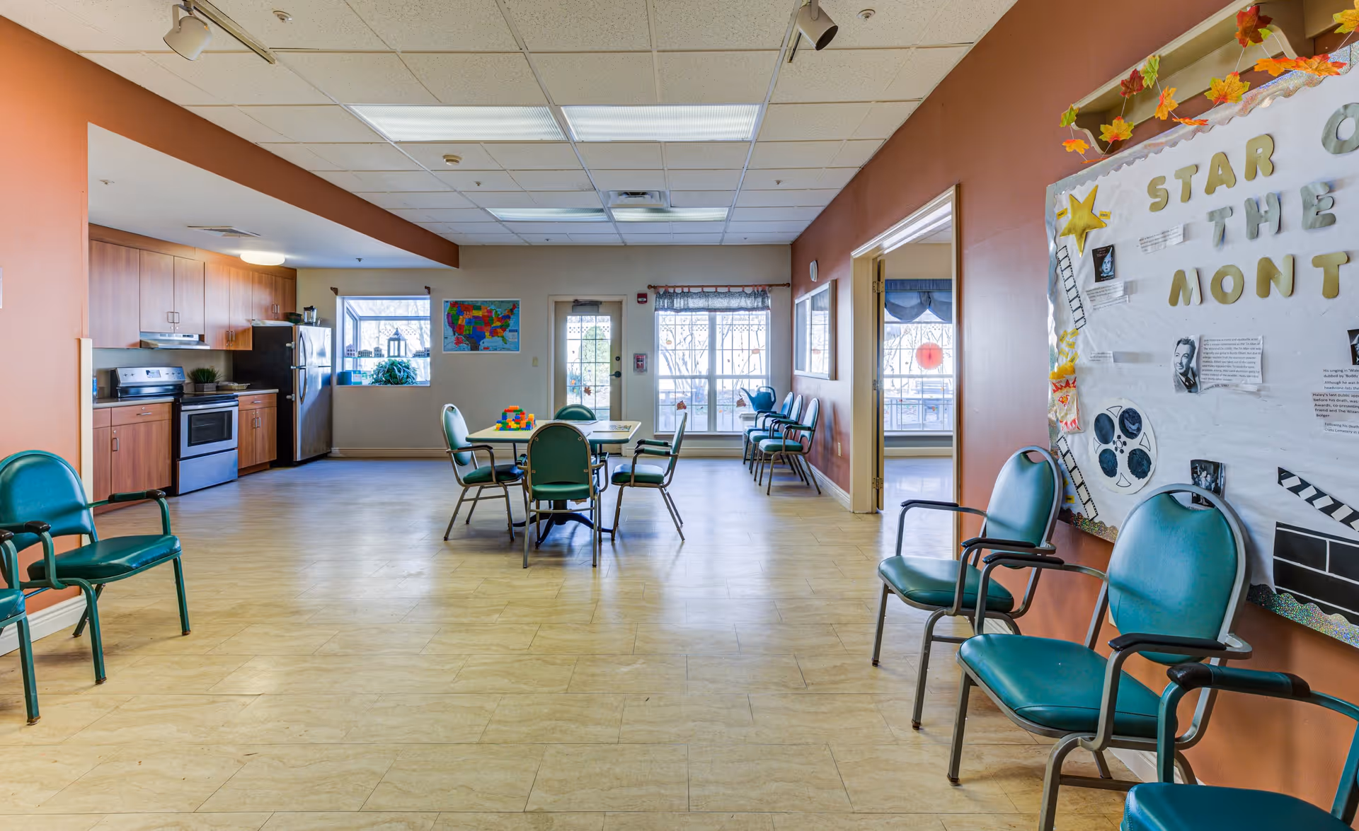 A spacious room with a kitchen area on the left featuring wooden cabinets, a stove, and a refrigerator. In the center, there are tables with green chairs around them. The walls are painted orange and beige, with a bulletin board on the right side displaying a 'Star of the Month' poster. Additional green chairs line the walls, and windows and a glass door at the back let in natural light.