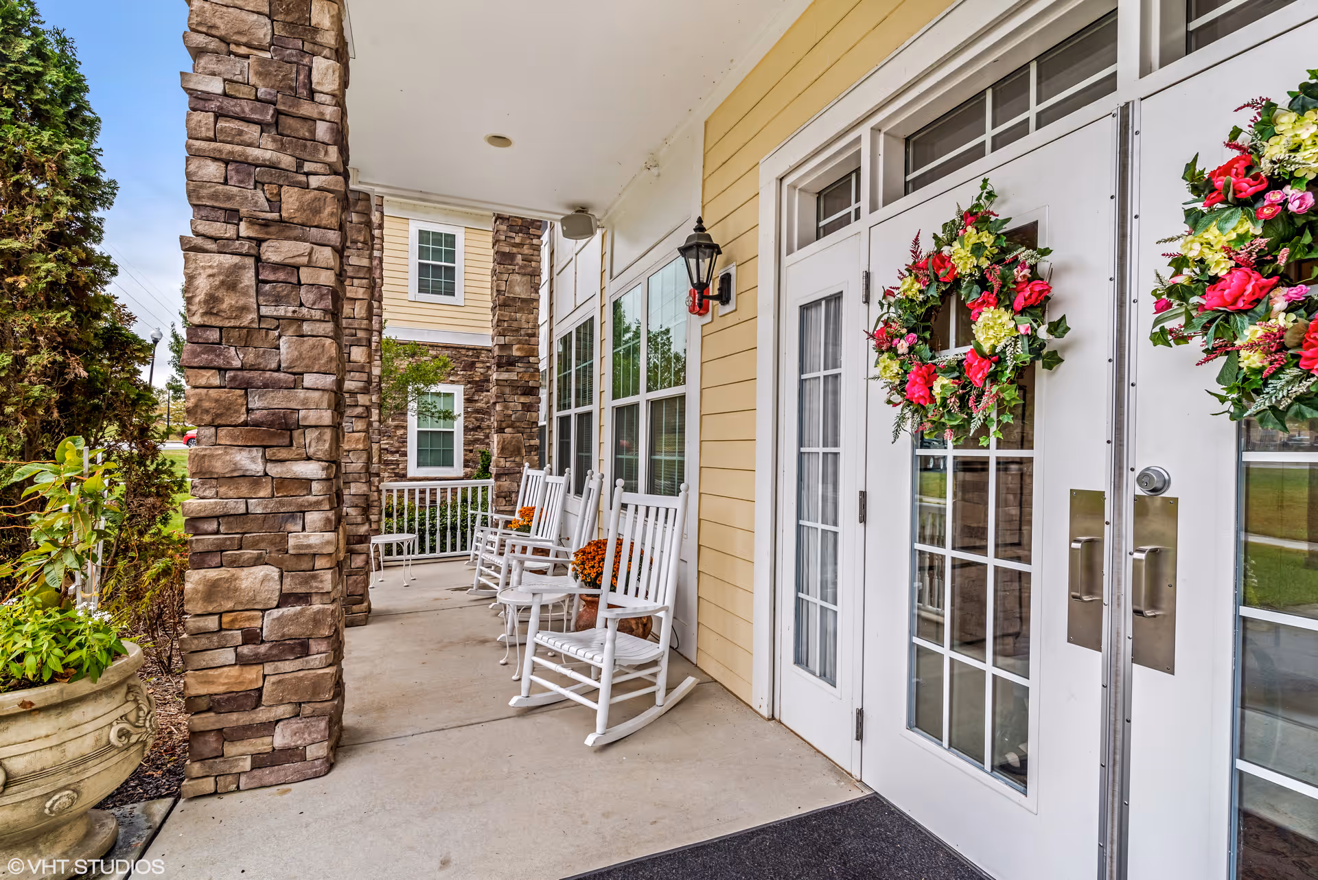 Covered porch area with stone pillars, white rocking chairs, and double glass doors decorated with floral wreaths. There are potted plants and a black outdoor wall lantern near the doors.