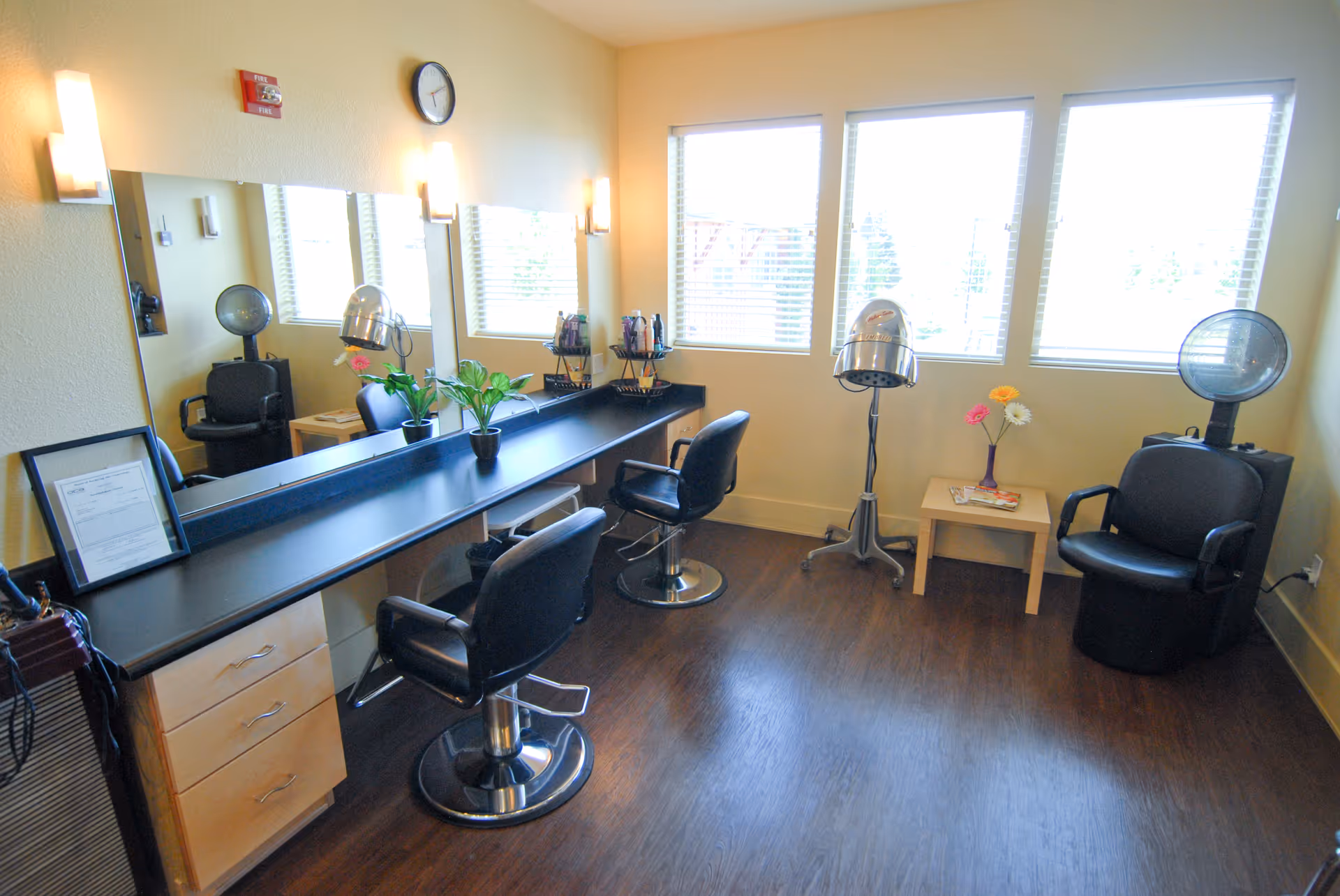 Interior view of a salon area in an assisted living facility with two black salon chairs in front of a long counter with a large mirror, two hair drying stations, a small table with flowers, and three large windows letting in natural light.