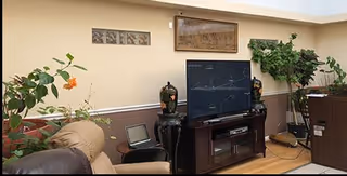 Interior view of a living room area in a senior living facility with a large flat-screen TV on a wooden TV stand, two decorative vases on either side of the TV, a reclining chair, a laptop on a side table, and several green plants along the walls.