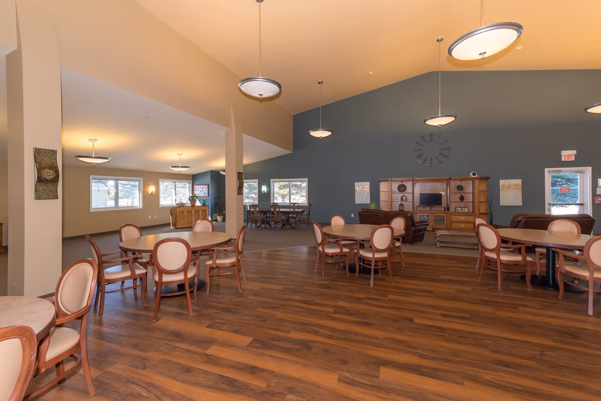 A spacious common area in a senior living facility with round tables and chairs arranged on a wooden floor. The room has a high ceiling with hanging light fixtures, a large clock on a blue-gray accent wall, and a wooden entertainment center with a TV. There are windows letting in natural light and a piano in the background near another seating area.