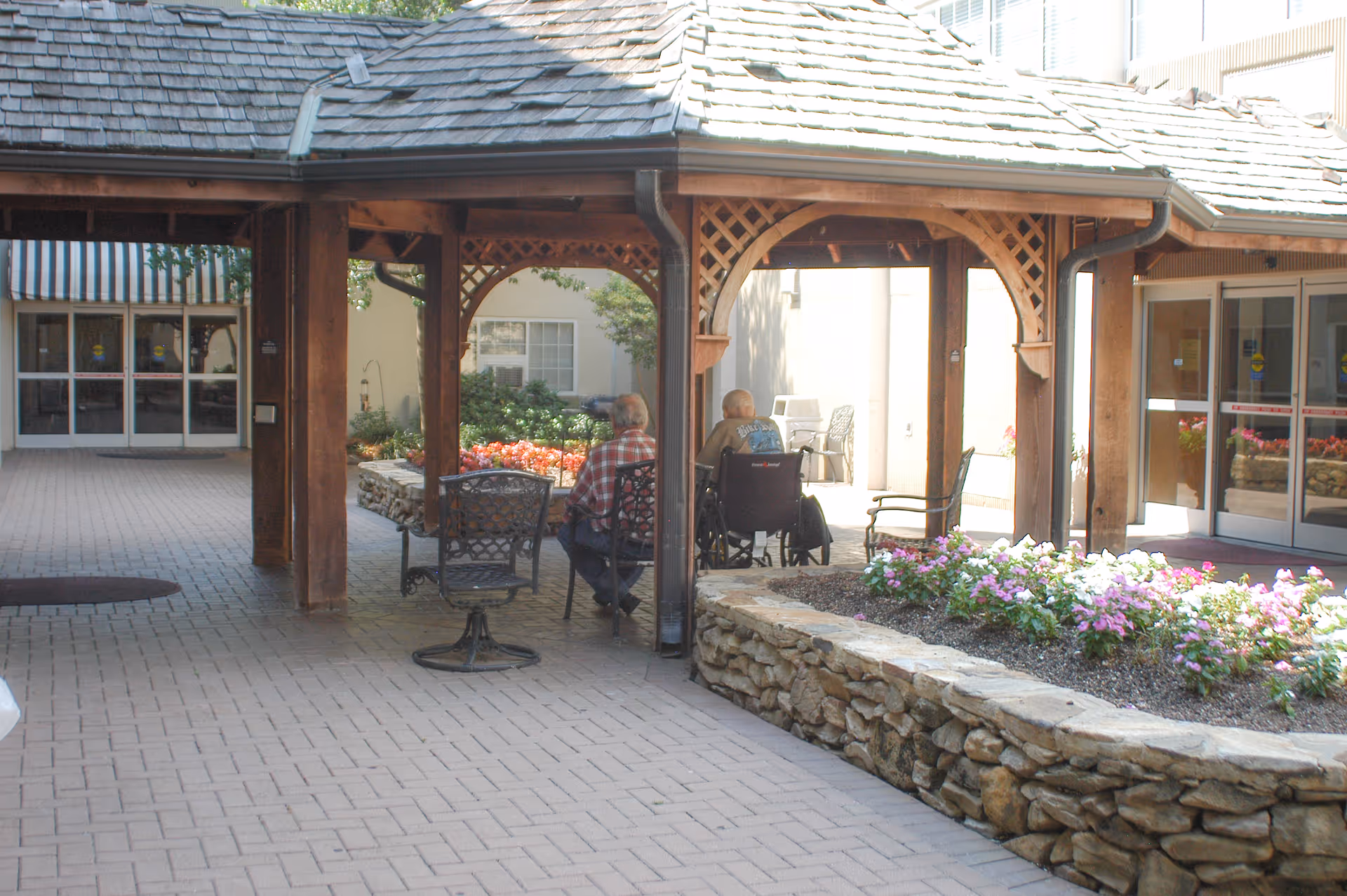 Covered outdoor courtyard at a senior living facility with seating, flower beds, and two residents under a wooden pergola.
