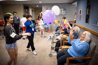 A group of elderly individuals seated in chairs and using walkers are playing with colorful balloons indoors, while younger people stand nearby engaging with them in a lively activity.