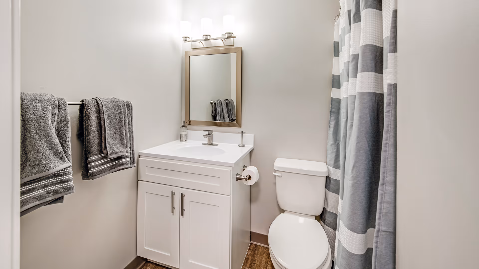 A clean and modern bathroom featuring a white vanity with a sink, a rectangular mirror above it, a toilet next to the vanity, a towel rack with two gray towels on the left wall, and a shower with a gray and white striped curtain on the right.