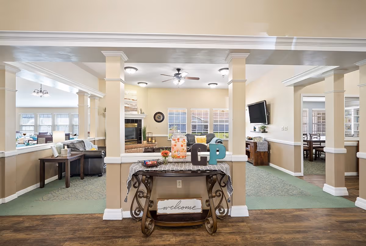 A bright and welcoming common area in a senior living facility with beige walls and green carpet. The room features a decorative table with a 'welcome' sign, a glass beverage dispenser, and decorative letters 'C' and 'P'. In the background, there is a seating area with sofas, a fireplace, a wall-mounted TV, and large windows letting in natural light. Adjacent rooms with dining tables and chairs are visible through open doorways.